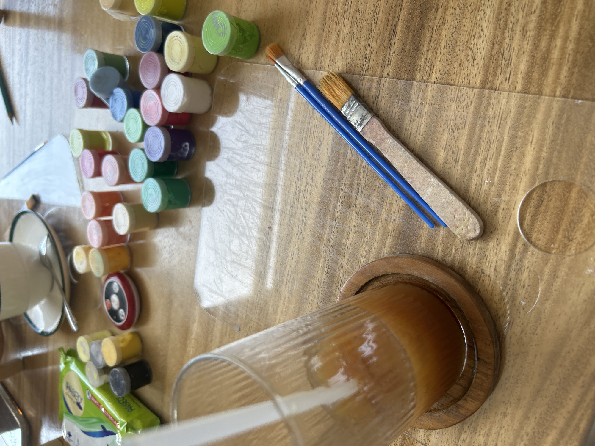 The scene is a tabletop shot of a collection of artistic and consumable items. The location is specified as Kuta Selatan, Indonesia.
Objects visible are: a variety of small, stacked paint pots in multiple colors; two paint brushes with blue handles; a glass of liquid (likely a beverage) resting on a wooden coaster with a straw inside; a sheet of clear plastic or acrylic; a cup and saucer with a spoon; and a package of wipes. The tabletop has a wood grain pattern. There are no people immediately visible in the frame. The items are arranged in a haphazard manner.