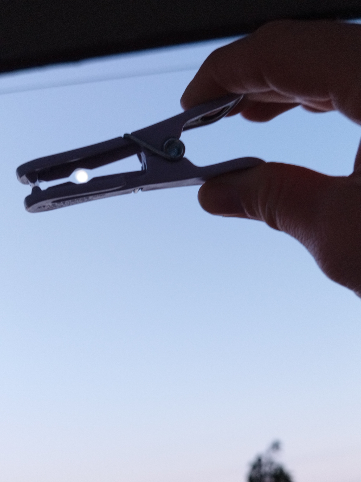 A hand holds a purple plastic clothespin in front of a gradient blue sky. The sun's light is visible through the open end of the clothespin. The scene is underexposed. The city of Sofia, Bulgaria is not identifiable in this image.