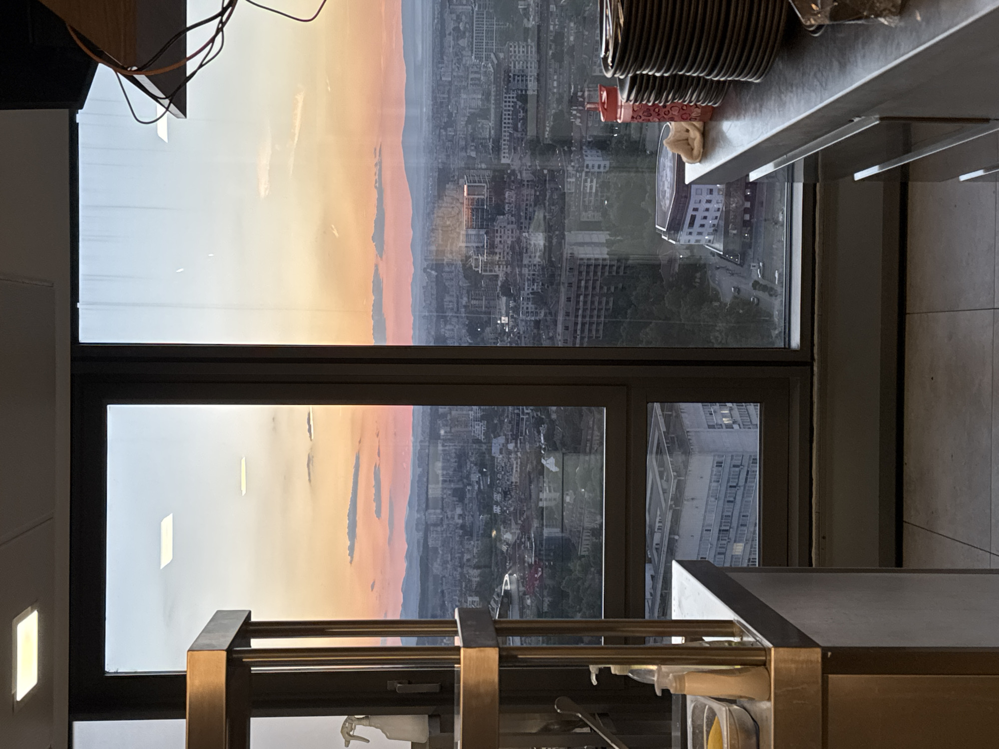 An outdoor panoramic view of the city of Sofia, Bulgaria, is captured through a large window during sunset. The sky displays a gradient of colors from pale blue at the top to orange and pink hues near the horizon, with distant mountains visible. The city below is densely populated with buildings of varying heights and architectural styles, reflecting the warm light of the setting sun. Trees and greenery are interspersed throughout the urban landscape.
In the foreground, the interior of what appears to be a commercial kitchen or prep area is visible. A stainless-steel shelving unit with two shelves is positioned on the left side of the frame. The top shelf holds a white spray bottle. The bottom shelf is partially obscured but contains a stainless-steel basin with yellow contents. Adjacent to this shelving unit is a stainless-steel countertop, extending to the right. On this countertop, a pump dispenser with white liquid and another clear bottle are present.
To the right of the shelving unit and on the countertop, a stack of dark-colored plates is neatly arranged. A red water bottle with a white heart pattern and a small folded white cloth are also on the countertop. The floor appears to be tiled. The overall scene suggests a functional space with a view, possibly a restaurant or catering establishment. The lighting indicates it is either early evening or late afternoon.