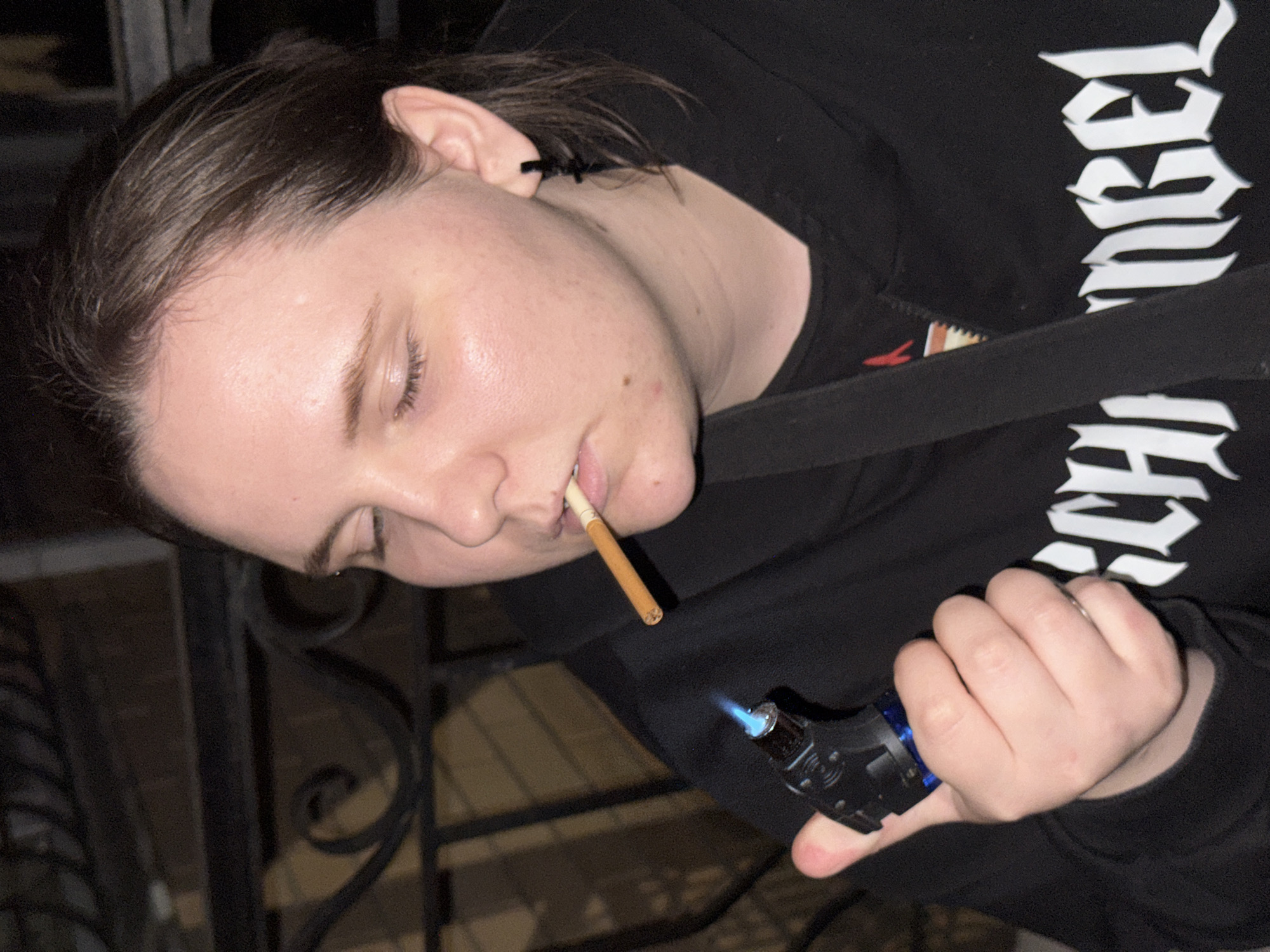 The image shows a close-up of a person, tilted on their side, with their eyes closed and a cigarette in their mouth. The person is wearing a black hoodie with white gothic-style lettering on the sleeve. In their right hand, they are holding a blue and black lighter, which appears to be lit, with a flame visible. The lighting suggests it is either nighttime or indoors in a dimly lit environment. The background is dark and indistinct, with hints of what might be a metal railing or fence. The overall mood appears relaxed or possibly weary, given the closed eyes. The visible text on the hoodie appears to be part of a word or phrase, with the letters "ngel" clearly visible.