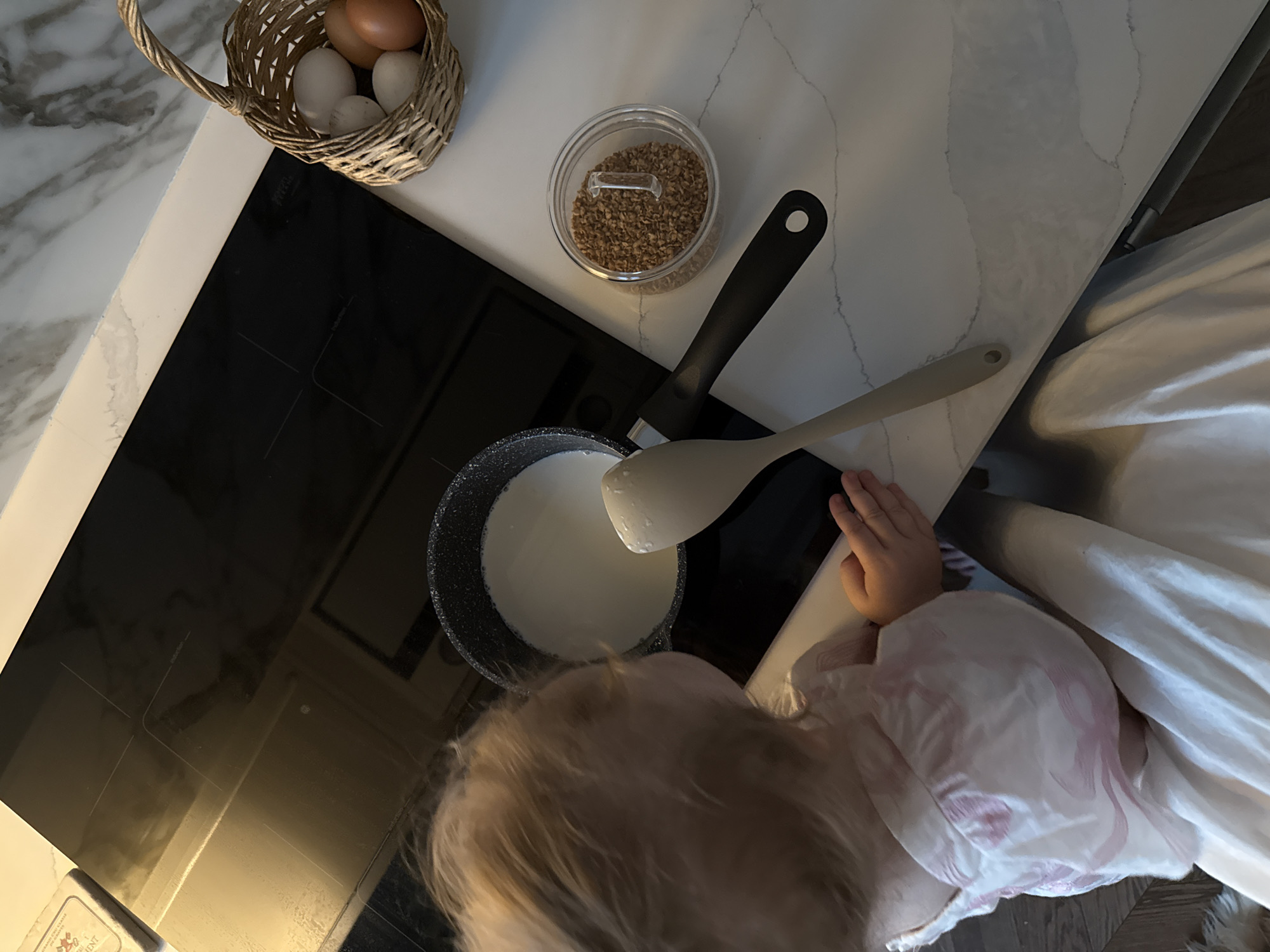 A top-down shot shows a kitchen scene where a small child is reaching towards a saucepan of milk on a black stovetop. A white spatula rests in the saucepan. To the right of the saucepan is a clear jar containing small brown pieces of food. To the upper left of the stovetop is a small basket containing eggs. A small tin is visible in the lower left corner of the stovetop. The scene is located on a countertop made of a light-colored material that may be marble. The child is wearing a light-colored, possibly patterned shirt or pajamas. The location of the image is not provided, and cannot be determined from the image itself. Moscow, Russia cannot be verified as the location, either.