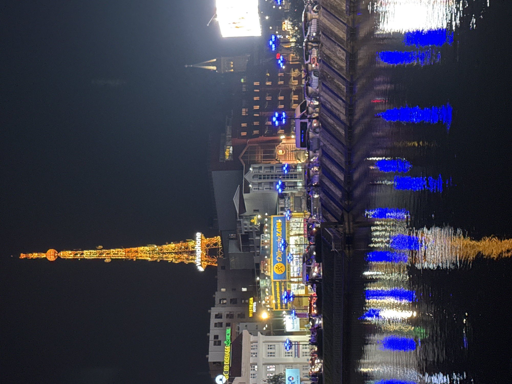 The image depicts a nighttime city scene, likely in Da Lat, Vietnam, characterized by illuminated buildings and a prominent tower. In the foreground, a body of water reflects the vibrant city lights, creating a colorful and somewhat distorted mirror image of the scene above. The water's surface is dark, serving as a canvas for the reflected blues, yellows, and whites of the urban illumination.
Dominating the skyline is a tall, slender tower, brightly lit with warm orange and yellow lights, giving it a metallic, skeletal appearance. A spherical light fixture sits atop the tower. Below the tower, various buildings are visible, some with illuminated windows and prominent signage. One sign, "vinaphone," is clearly visible on the tower structure.
Further down, a street level view shows a bustling environment with a line of cars and motorcycles. Blue lights from street lamps or decorative elements are prominent, casting a cool glow on the scene and contributing to the reflections in the water. Buildings at this level are also illuminated, with some displaying shop signs. One sign appears to read "Điện máy XANH" and another "GOLD DREAM." The architecture suggests a blend of modern and older structures. A church steeple is visible in the background to the right, also lit. The overall impression is one of an active urban area at night, with a strong emphasis on artificial lighting. There are no discernible individuals clearly captured in motion or interaction.