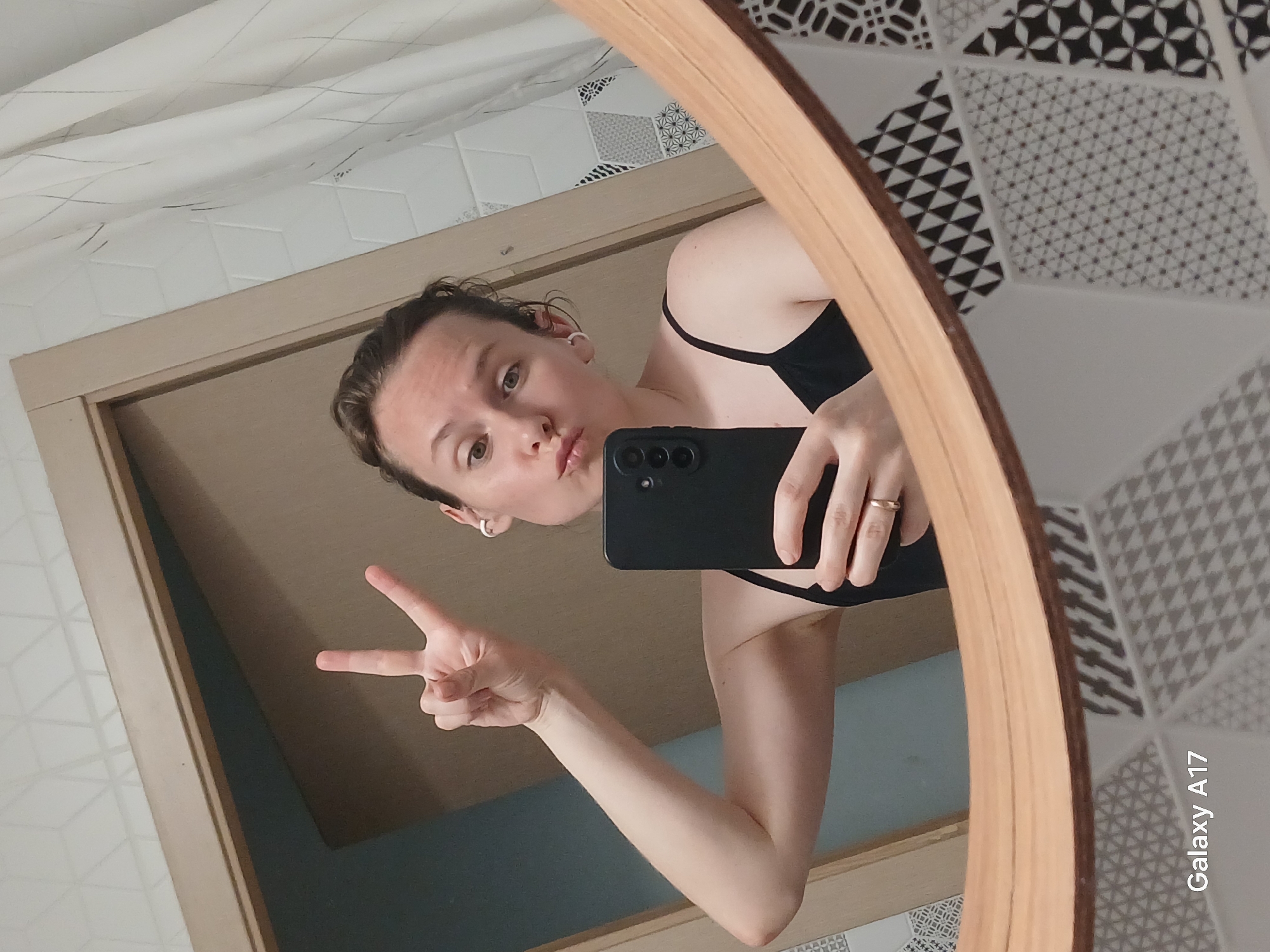 A selfie taken in a mirror, showing a fair-skinned person with short brown hair, wearing a black top, holding a black smartphone with a visible camera array in their right hand and making a peace sign with their left hand. The person is making a kissing face. The mirror is round and framed with wood. The background shows geometric-patterned wall tiles and a shower curtain or a textured wall covering. "Galaxy A17" is visible in the lower right corner. The location appears to be a bathroom, possibly in a residential building. It is impossible to determine if the image was taken in Yekaterinburg, Russia, without additional information.