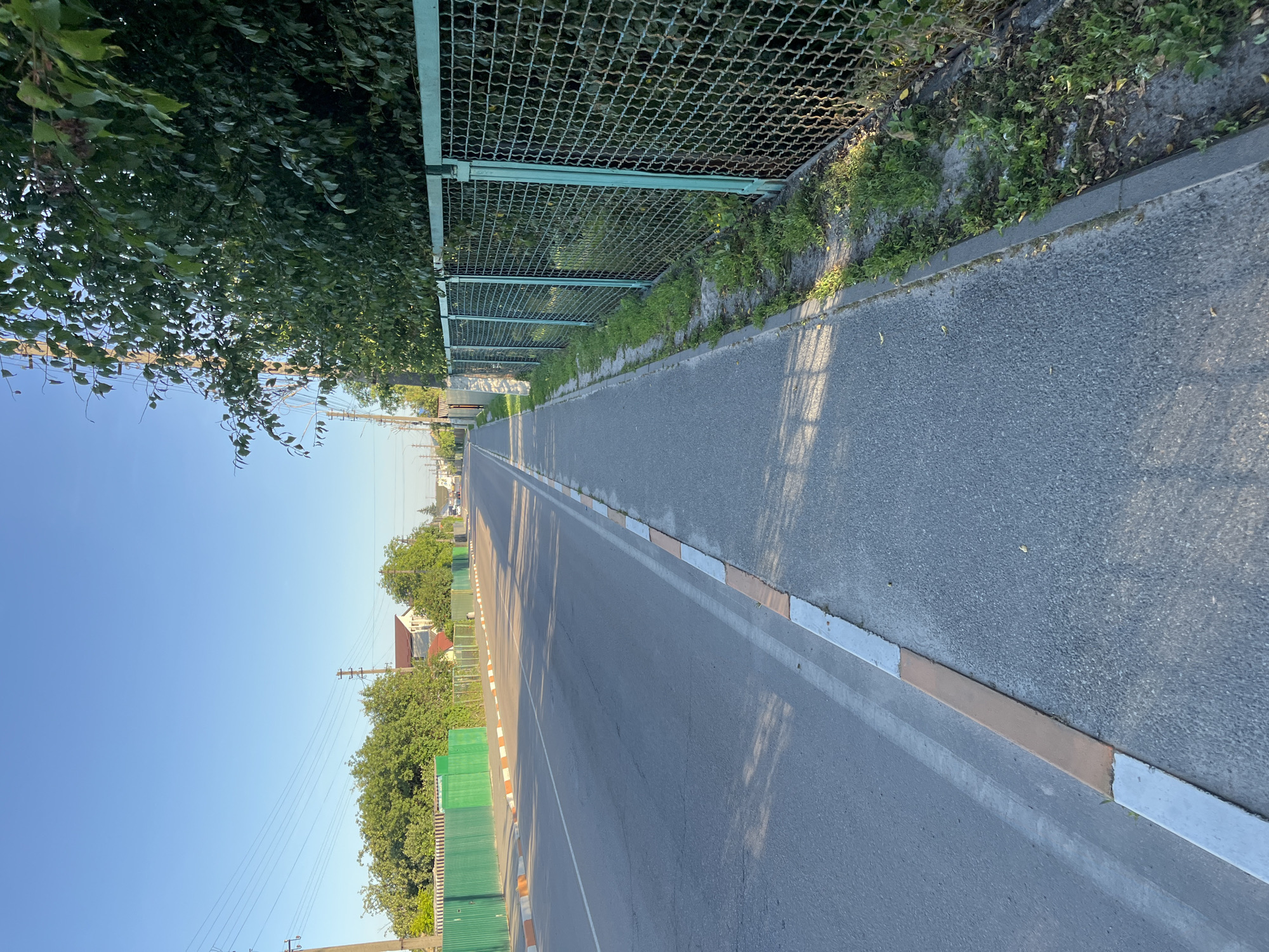 This is a vertical, eye-level, outdoor photograph taken on a sunny day in the city of Kalynivka, Ukraine. The image captures a view of a paved road with a sidewalk running alongside it. The road has a white dashed line marking the center and a white solid line along its right edge. The sidewalk is bordered by a curb with alternating orange and white painted sections. To the right of the sidewalk is a green metal mesh fence, beyond which dense green foliage is visible. In the distance, houses with red roofs and trees can be seen. Power lines are strung across the sky. The overall scene depicts a quiet, residential street.