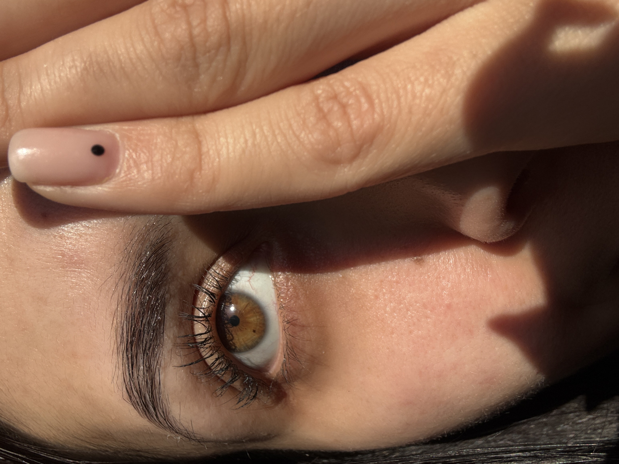 A close-up, angled shot focuses on a person's face, with their hand partially covering their eyes. The visible eye is brown, surrounded by dark eyelashes and eyebrow. The skin is fair, and a fingernail with a single black dot design is visible. The background is dark and blurry. The location, Molodezhnyi, Russia, is not discernible from the image.