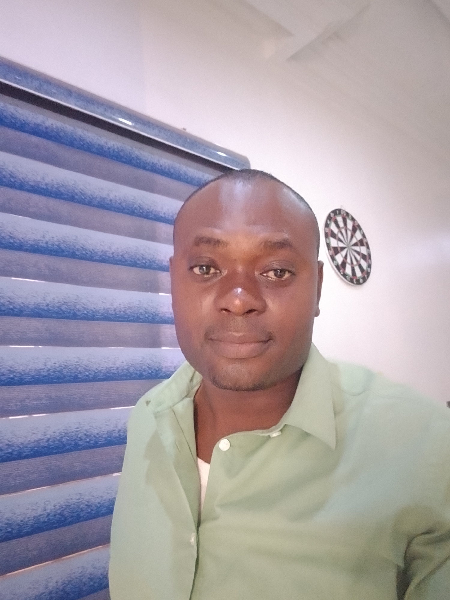 A head and shoulders shot of a man in Gwarinpa, Nigeria. He has dark skin, short hair, and is wearing a light green collared shirt over a white undershirt. Behind him is a window with closed horizontal blinds of alternating blue and white stripes. Further back, on the white wall, is a dartboard. The lighting is somewhat dim.
