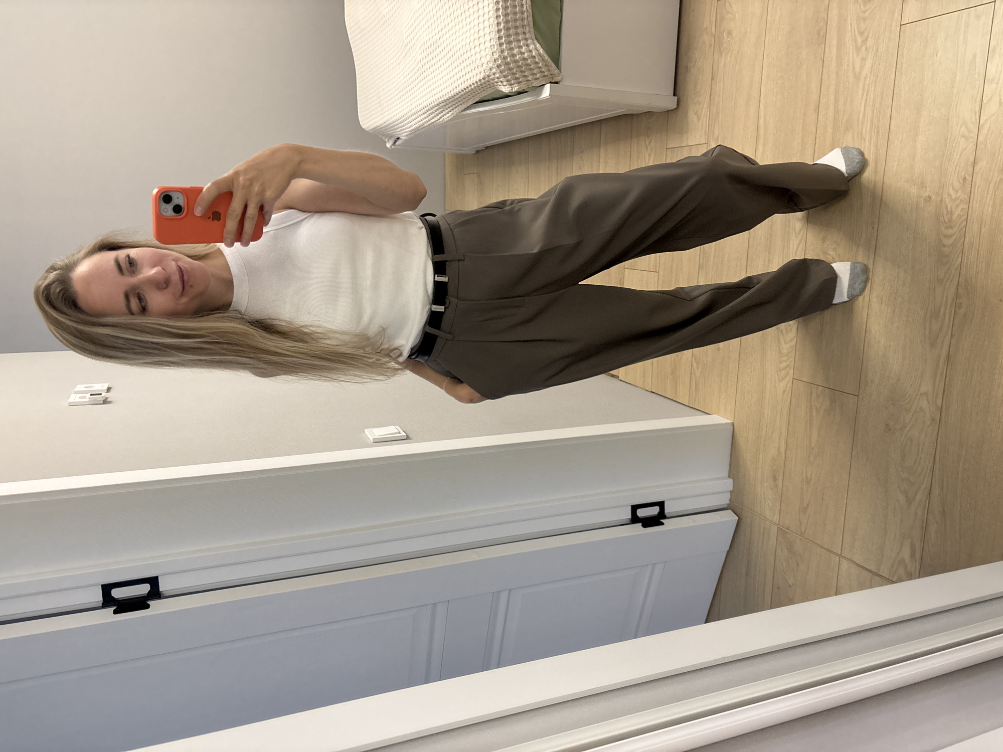 A woman with long blonde hair is taking a mirror selfie in a room. She is wearing a white sleeveless top, brown wide-leg pants, a black belt, and grey and white socks. She is holding an orange smartphone in her right hand. The room has light beige walls, a wooden floor, a white door with black hardware, and a white bed with a beige textured blanket. The location is an indoor residential setting in the city of Chaikovsky, Russia.
