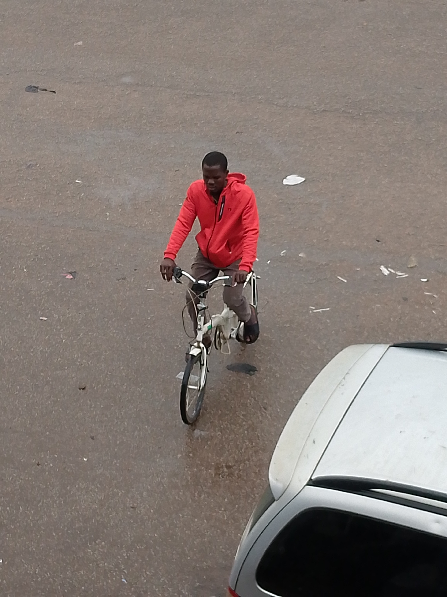 An overhead shot captures a man riding a white bicycle on a paved road in Bukuru, Nigeria. The man, with dark skin, is wearing a vibrant red hooded jacket, dark trousers or shorts, and black flip-flops. He is positioned centrally, holding the bicycle's handlebars, and appears focused on his path, looking slightly downwards. The bicycle has a white frame and visible spokes on its front wheel.
In the lower right corner, the rear section of a silver or light-colored car is visible, featuring a spoiler. The road surface beneath them is a light grey asphalt, showing signs of dampness or scattered dirt and small pieces of debris. The lighting suggests it is daytime, possibly overcast, as there are no distinct shadows. No visible text can be discerned on any of the subjects or objects in the image. The scene depicts a commonplace urban or semi-urban activity.