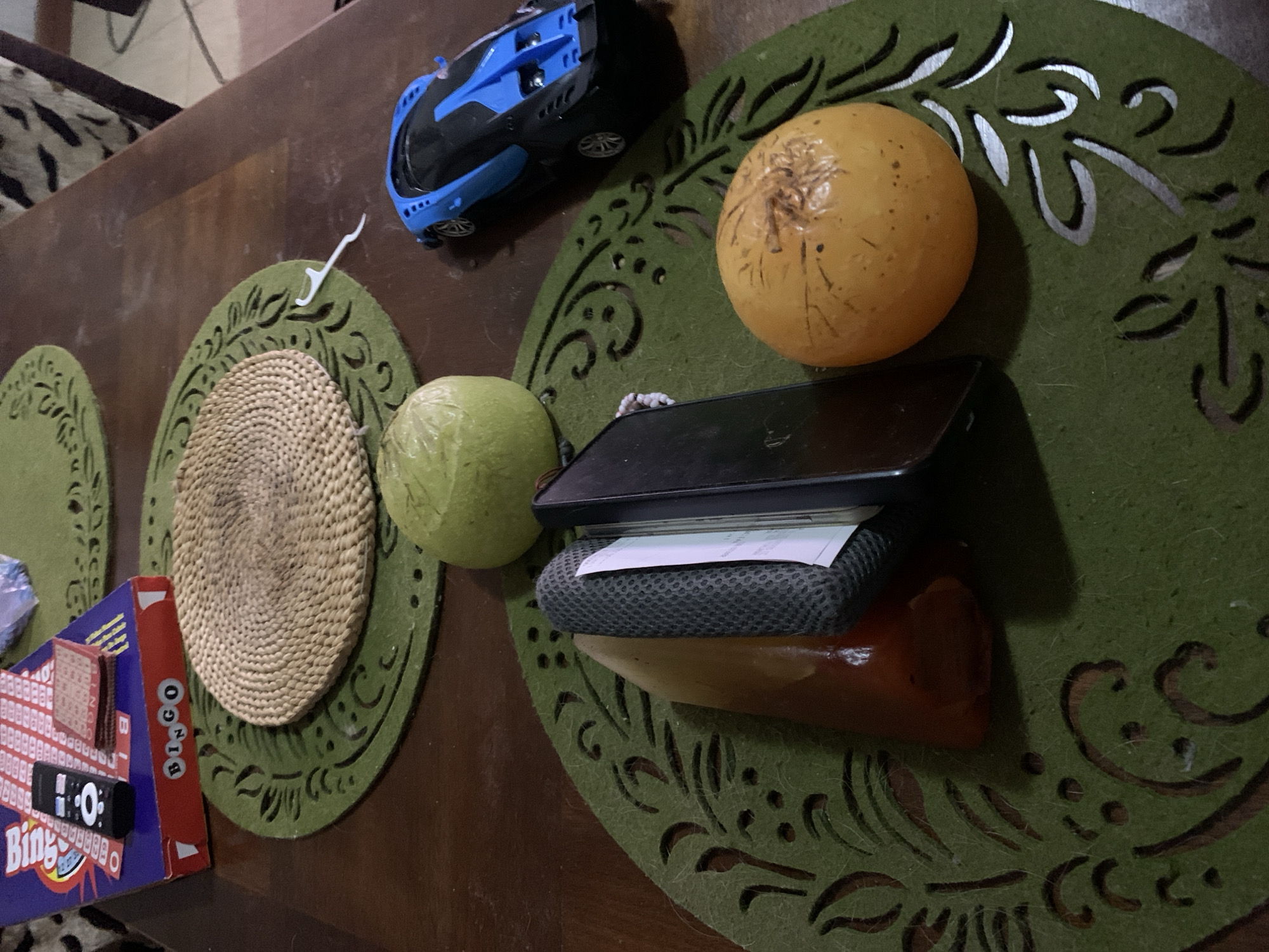 The image shows a collection of items on a dark wooden table. There are three green, circular placemats with decorative cutouts. Clockwise from the left, objects include a Bingo game box, a woven placemat, a green guava, a blue toy car, an orange fruit, a black cell phone on top of a stack of items including a gray mesh item, a white paper, and an orange item. The scene appears to be indoors. There is no information in the image to identify the location in Santa Ana, Venezuela.