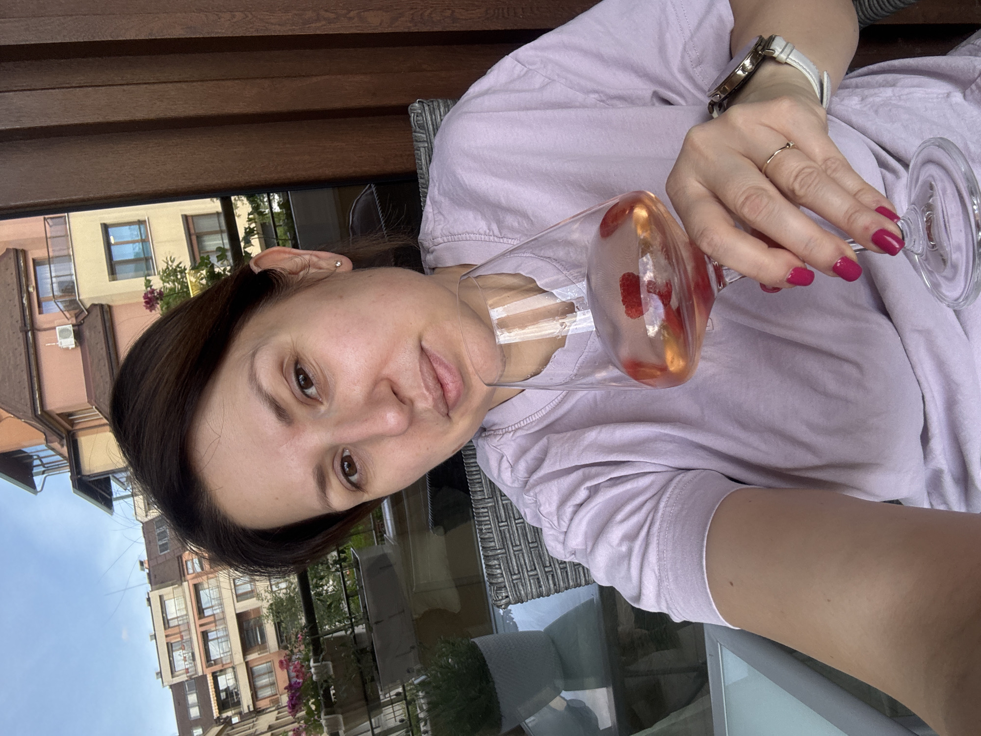 A woman is holding a glass of light pink liquid, potentially wine or juice, with raspberries visible inside the glass. She is wearing a white watch with a gold bezel and a thin gold ring on her finger. Her nails are painted a dark pink/red color. She is wearing a light purple t-shirt. The background shows a building with multiple stories, and the overall scene suggests an outdoor location, possibly a balcony or patio in the city of Sofia, Bulgaria. The time of day appears to be daytime, evidenced by the natural light.