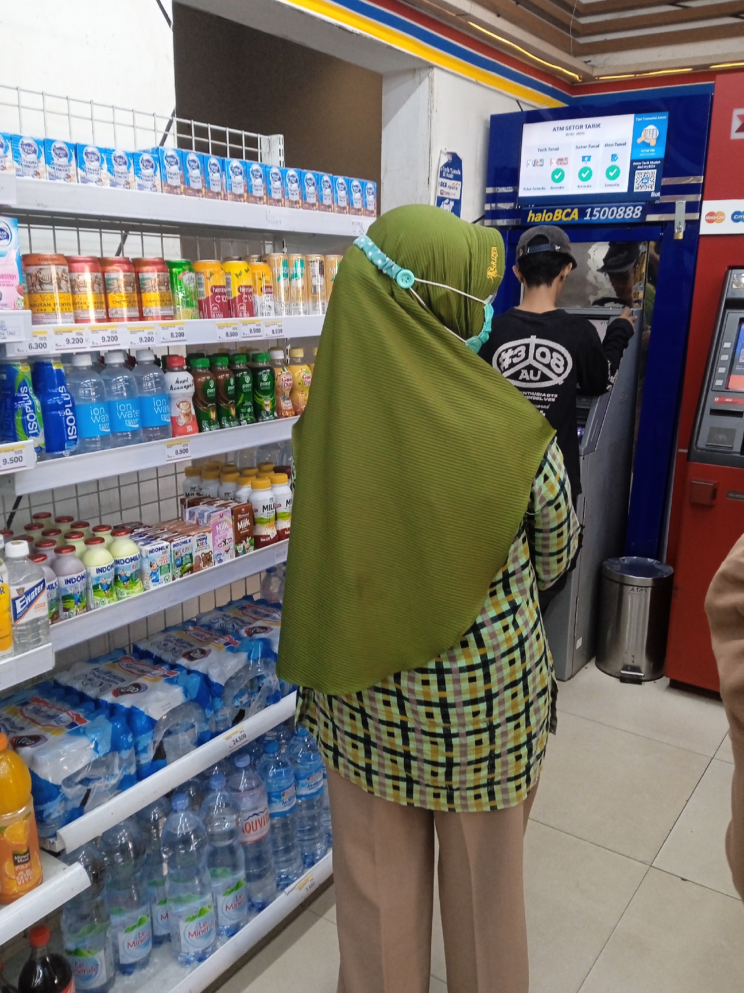 In Kaliwungu, Indonesia, inside a convenience store, a woman wearing a green hijab and a plaid shirt stands in front of an ATM. She has a surgical mask on, held in place by a light blue beaded strap. To her right, a young man in a black t-shirt with white lettering and a baseball cap is using the ATM. The ATM screen displays transaction options and the phrase "haloBCA 1500888." Shelves filled with beverages, including bottled water, milk cartons, and canned drinks, are visible in the background. A metal trash can is positioned to the right of the ATM. The floor is tiled.