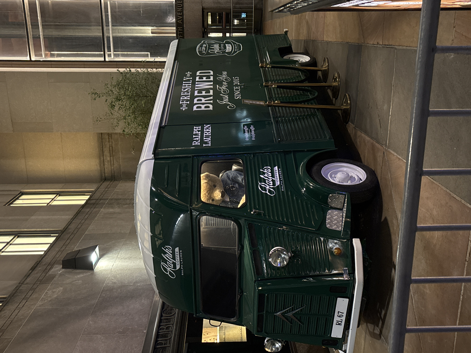 An outdoor, nighttime shot captures a dark green vintage-style coffee truck, likely a modified Citroën Type H van, illuminated by artificial light on what appears to be a city sidewalk. The truck is prominently branded "Ralph's Coffee" by Ralph Lauren, with large white text on its side reading "FRESHLY BREWED Just For You SINCE 2015." Other branding includes "@ralphscoffee" and "#ralphscoffee." In the driver's seat, a light brown teddy bear is visible, adding a whimsical touch.
On the passenger side, three gold-colored stanchions are set up, suggesting a service area or queue line. The front of the truck displays a "RL 67" license plate and the distinctive Citroën chevron logo. The vehicle's wheels are white, contrasting with its dark green body.
The truck is parked alongside a tall building with large windows, some of which are lit. Part of a street sign reading "FIFTH AVENUE" is visible in the upper background, placing the scene in a prominent urban location, specifically New York City, United States of America. A street lamp casts light on the building wall near the front of the truck. The ground is paved, and a metal railing is partially visible on the right side of the frame. The overall atmosphere is inviting and stylish, indicative of a premium mobile coffee service operating in an upscale city environment during the evening or night. Additional visible text includes "A CUP OF COFFEE" near the roofline and a partially visible "PUBLIC" sign on the building.