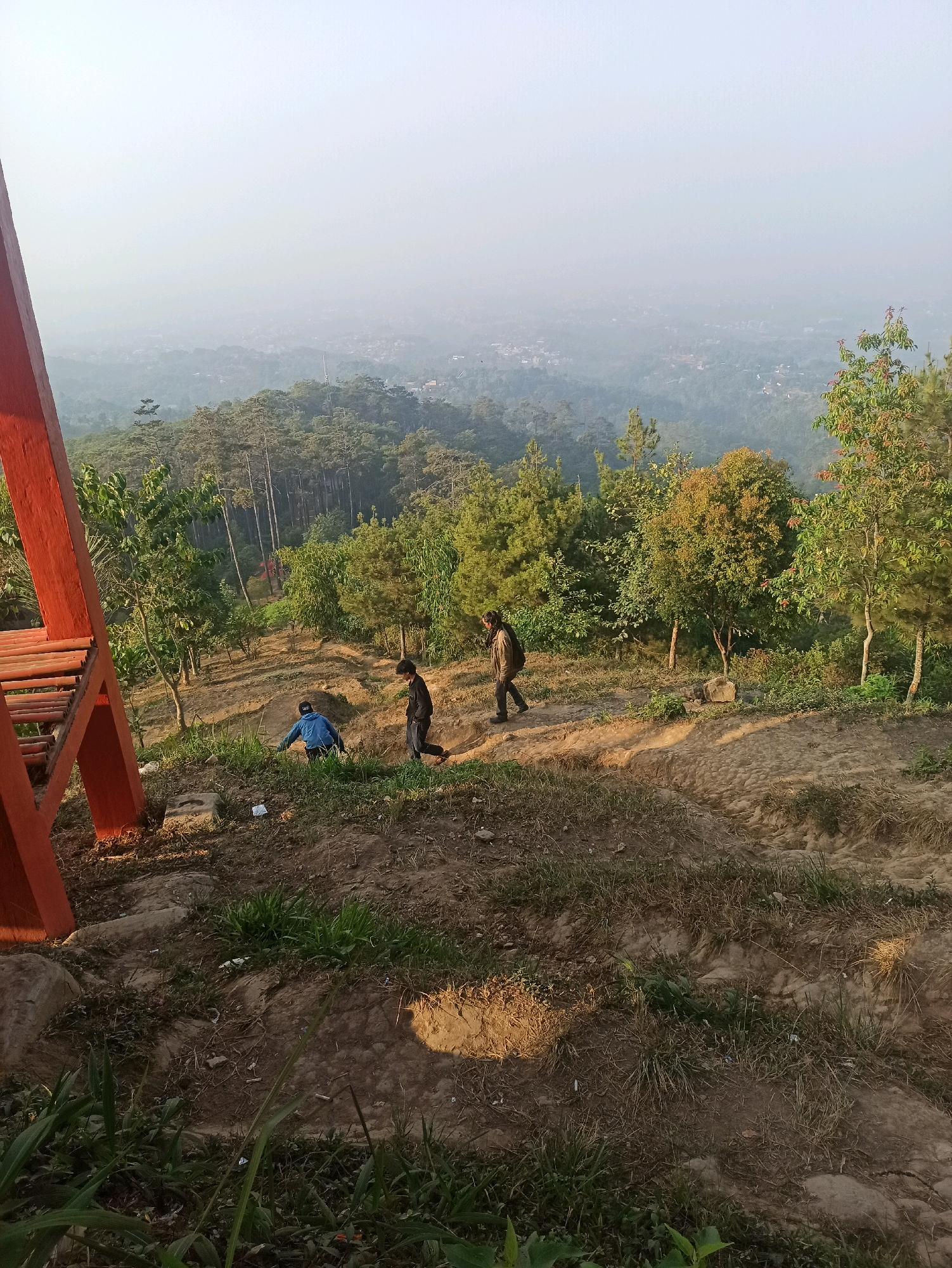 In Cileunyi, Indonesia, an outdoor scene depicts a hazy, elevated landscape viewed from a slightly sloped, dirt path. The foreground features a large, bright orange wooden structure on the left, with visible bamboo planks. Three individuals are present on the path. To the left, a person in a blue jacket is crouched down. Further up the path, two individuals are walking away from the viewer; one in a black jacket and dark pants, the other in a brown jacket and dark pants with a backpack. The middle ground is a wooded area with various trees, including tall pines and deciduous trees with some autumnal foliage. The background reveals a sprawling city nestled in a valley, obscured by a thick haze that stretches to the horizon. The ground is a mix of dry soil, sparse grass, and scattered debris.