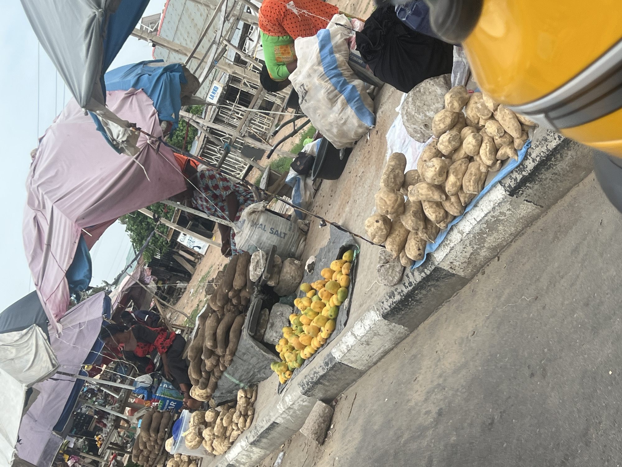 The image depicts a street scene in Jalingo, Nigeria, showcasing a bustling market environment. Numerous vendors display their wares along the roadside under makeshift shelters constructed with tarps and fabric. Piles of yams, mangoes, and sweet potatoes are prominently featured, offered for sale on the edge of the roadway. People are present, presumably as vendors and potential customers, positioned amidst the market activity. Bags of goods, including what appears to be salt and cement, are also visible. A vehicle is partially visible in the foreground. The general atmosphere is one of active commercial exchange.