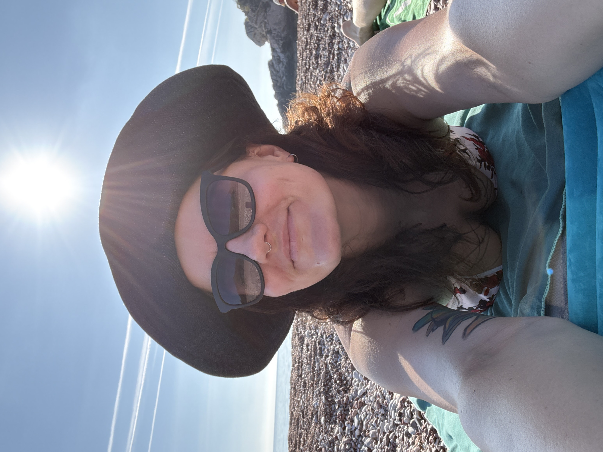 A woman wearing a black hat and sunglasses is lying on a beach. The beach is covered in small pebbles. The woman is wearing a floral bikini top and is covered by a light green and blue blanket. There is a tattoo on her left arm. The sun is shining brightly. In the background, there is a rocky cliff and the sea. The location appears to be Sveti Stefan, Montenegro.
