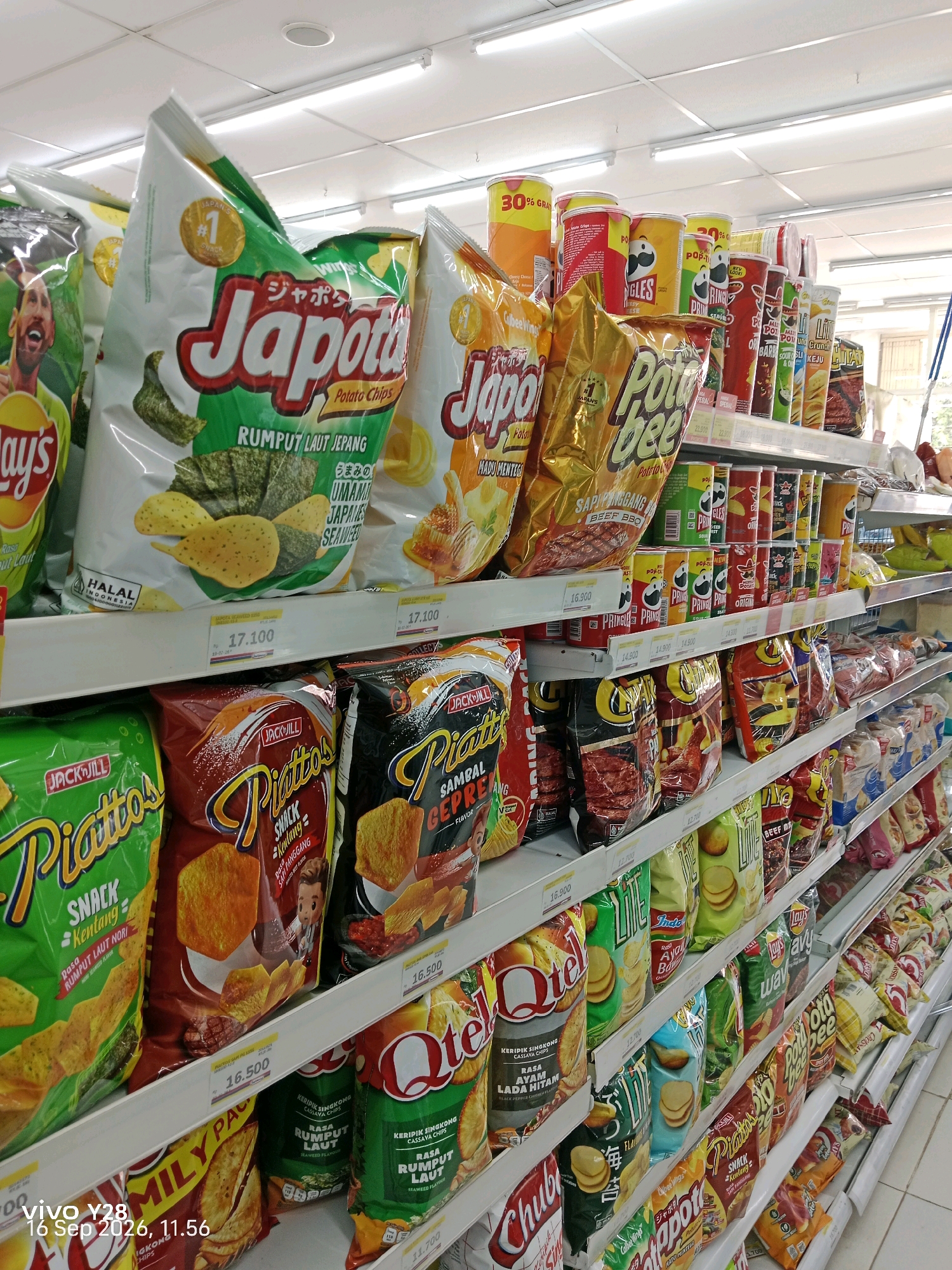 This image is taken inside a convenience store in Tlogosari, Indonesia, likely a minimarket. The perspective is from eye-level, looking down an aisle filled with shelves stocked with a variety of snack food products. The products are primarily potato chips and similar packaged snacks, arranged in bags and cylindrical containers. Dominant brands visible include Japota, Pringles, Piattos, and Lay's. The shelves are white, and the lighting appears to be fluorescent, typical of a retail environment. The flooring is a light-colored tile. The image is cropped to focus on the snack products, with no people visible in the frame. The composition suggests a consumer's view of a well-stocked snack aisle, possibly for the purpose of product cataloging or retail display analysis. The timestamp "vivo Y28 16 Sep 2026, 11.56" in the lower left corner is likely a watermark from the camera or phone used to capture the image.