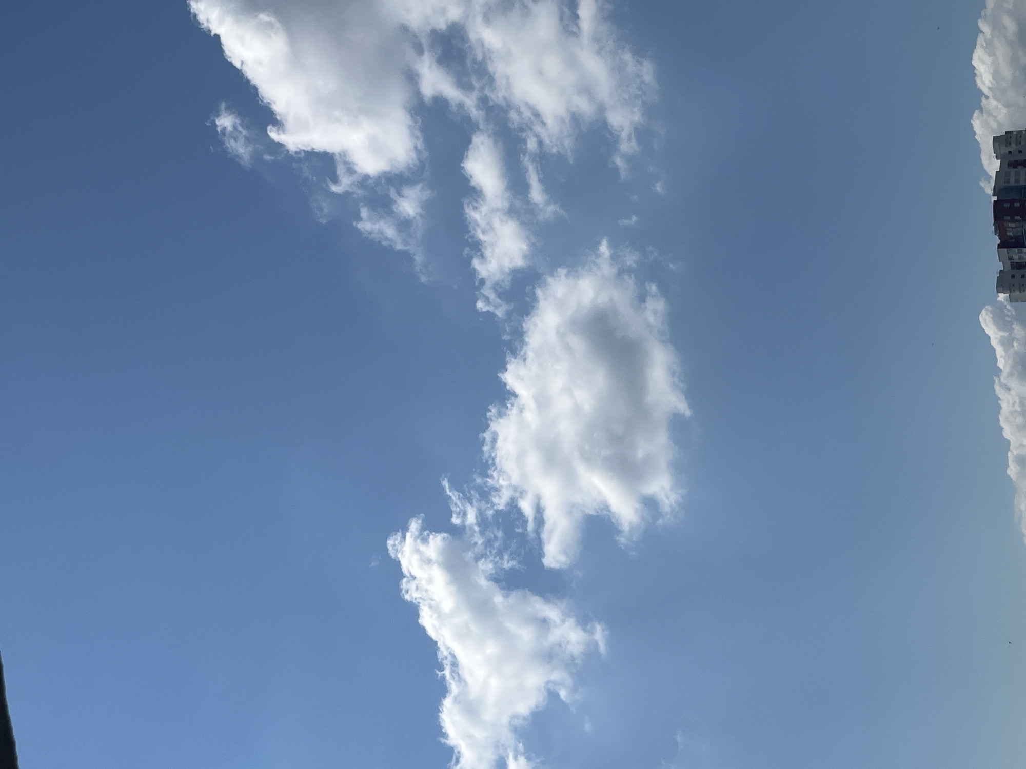 This image shows a blue sky with scattered white clouds. In the lower portion of the image, the tops of buildings are visible. One building in the center appears to be a residential complex with red and white sections. The clouds are wispy and appear to be cumulus clouds. The lighting suggests it is daytime with bright sunlight.