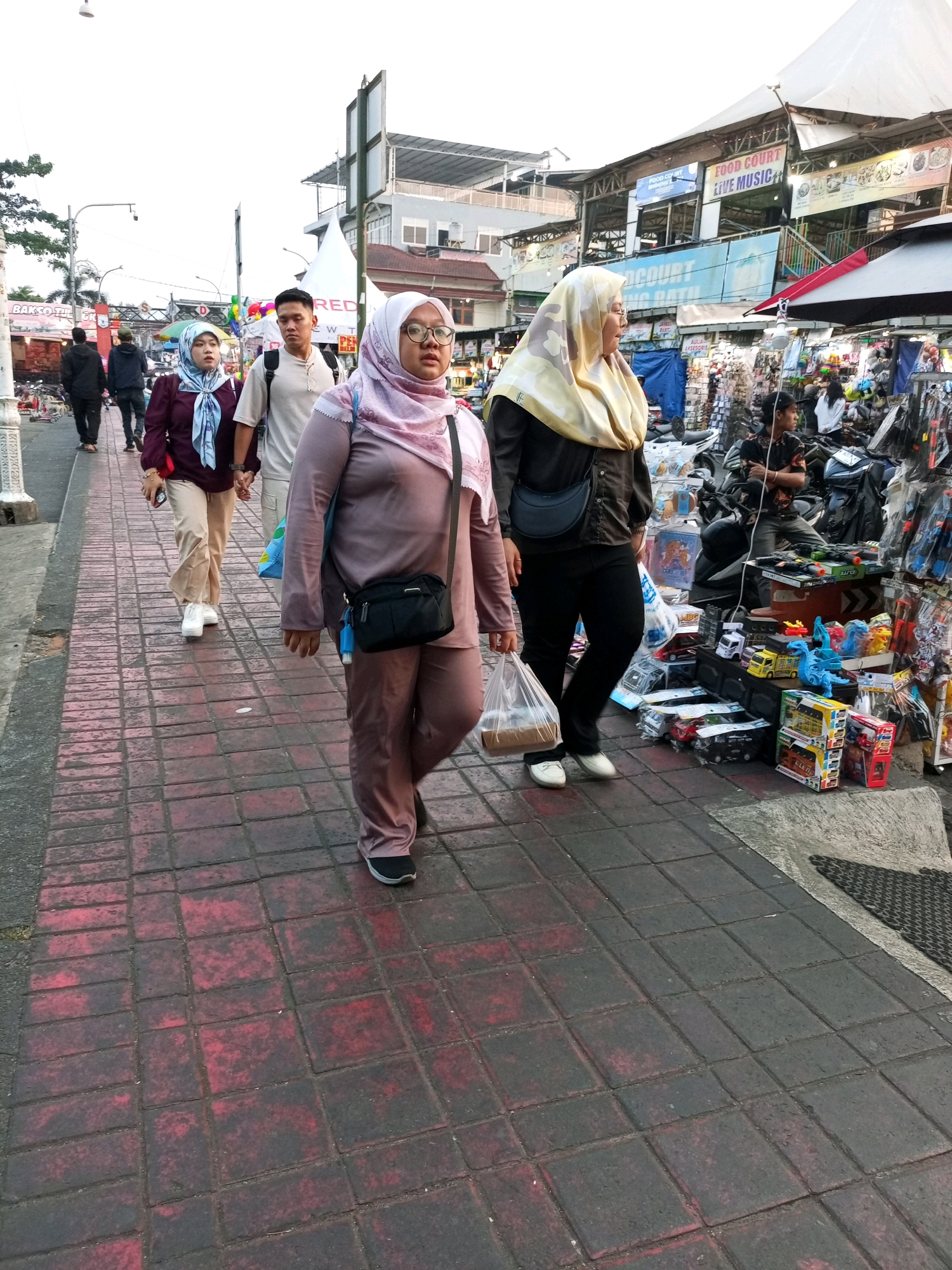 In the city of Batu, Indonesia, this image captures a street scene where several individuals are walking along a red-tiled pedestrian walkway. In the foreground, a woman wearing a pink headscarf and matching pantsuit is walking towards the viewer, carrying a plastic bag in her right hand and a black crossbody bag. To her left, a couple is walking away from the viewer; the woman wears a maroon top and light-colored pants, while the man in front of her wears a light-colored t-shirt. Further in the background, other pedestrians are visible, including two men in dark clothing walking away from the camera. On the right side of the walkway, various stalls are set up, displaying goods such as toys and accessories. Buildings with signage, including "FOOD COURT LIVE MUSIC", are visible behind the stalls. The overall atmosphere suggests a bustling market or street-side shopping area.