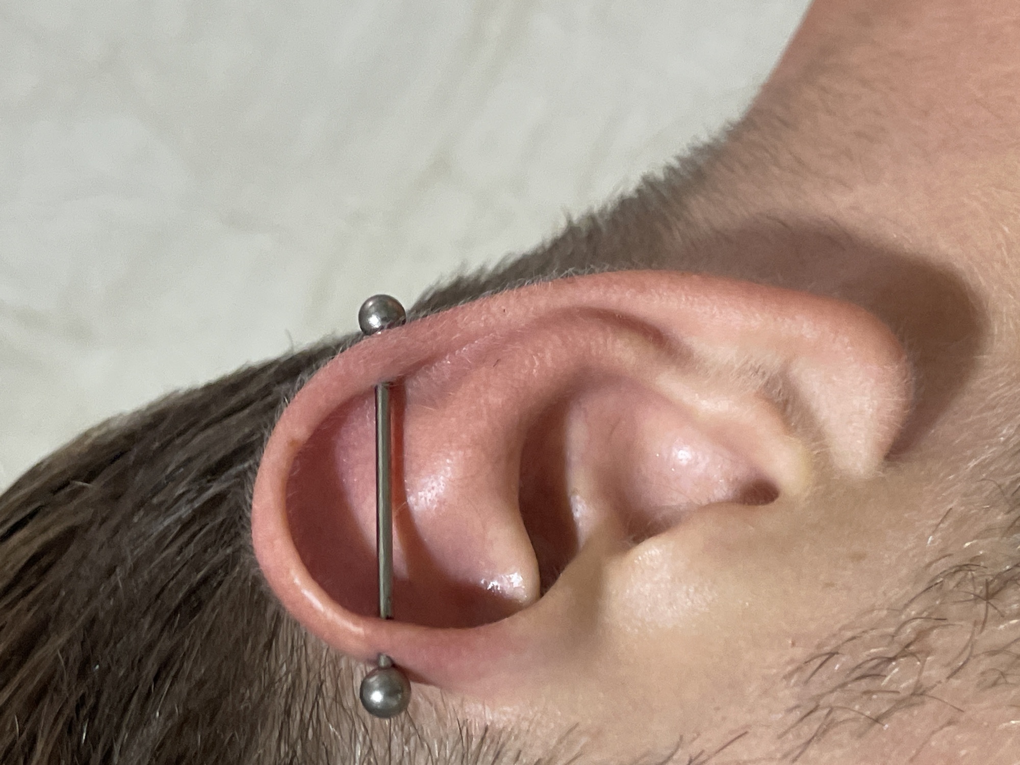A close-up, profile view of a person's ear with an industrial piercing. The piercing consists of a silver-toned barbell that passes through two points on the upper cartilage of the ear. The barbell has a spherical bead at each end. The skin of the ear is pink and appears smooth. The person has short, dark brown hair that is slightly wet. The background is a plain, light-colored wall. There is no visible indication of the location being Hoi An, Vietnam.