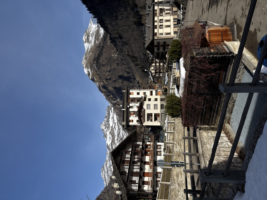 The image presents a panoramic view of a mountain village or town nestled beneath towering, snow-capped peaks under a clear, bright blue sky. The scene is captured during daytime, with direct sunlight illuminating the landscape and casting distinct shadows, suggesting either morning or early afternoon.
In the foreground and mid-ground, a collection of buildings of varying architectural styles forms the core of the settlement. To the far right, a wooden railing runs along what appears to be a path or elevated area. Behind it, a rustic wooden structure, possibly a shed or covered walkway, is visible, partially overgrown with reddish-brown, bare thorny bushes. A patch of snow is visible on the ground in the immediate foreground, indicating winter or early spring.
Towards the center-right, a multi-story building with a light-colored facade and numerous windows stands prominently. Further left, another multi-story building with a similar light-colored exterior features several satellite dishes on its roof or upper exterior walls, and some windows have dark shutters. To its left, a larger, more traditional-looking building with dark wooden walls and multiple stories, possibly featuring balconies or open-air corridors, dominates that section of the village. Scattered throughout the village are other buildings, some with light facades and dark roofs, blending into the mountainous backdrop. Stone walls and terracing are also visible, hinting at the challenging terrain.
The village is situated at the base of dramatic mountains. In the background, rugged, dark mountains rise majestically, with significant patches of snow clinging to their higher elevations, particularly on the prominent peak in the upper right. Further to the left, additional snow-capped peaks are visible against the clear sky. The lower slopes of the mountains are covered with a dense growth of dark, bare trees, transitioning into the built environment of the village. The bright blue sky fills the upper portions of the image, accentuating the clarity of the day.
No specific activity or individuals are visible, rendering the scene calm and tranquil. There is no visible text in the image.