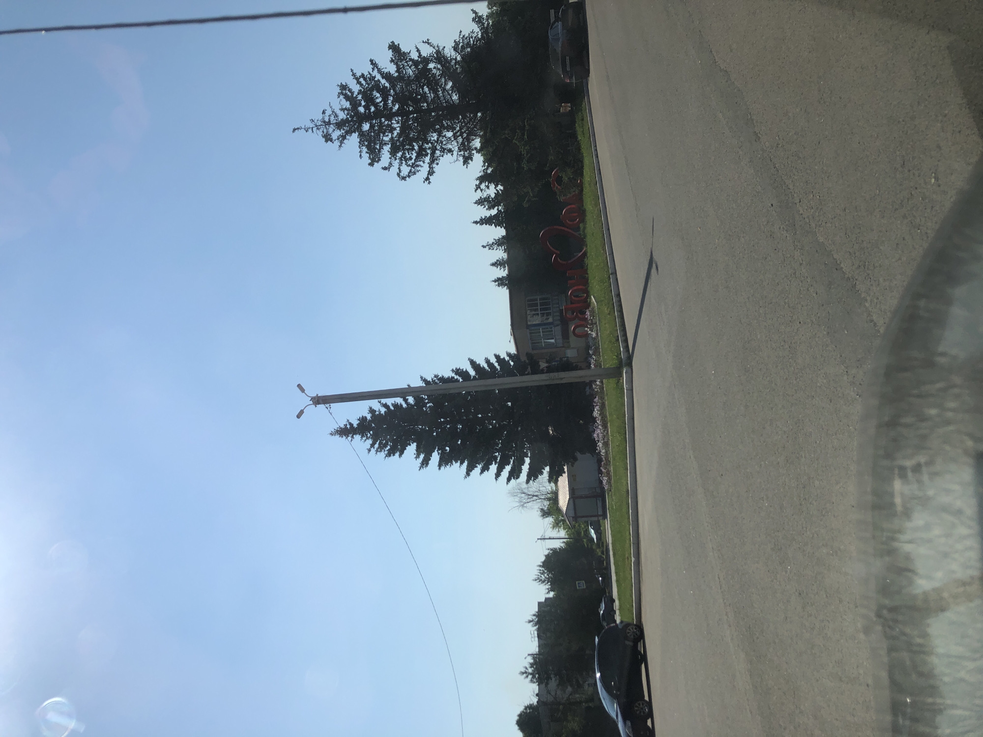 In Maksimovo, Russia, on a clear, sunny day, a street view shows a large Christmas tree in the foreground with a lamppost nearby. In the background, red decorative letters spelling "MAKSIMOVO" with a heart are visible in front of a building surrounded by trees. A dark car is parked on the street to the right of the letters, and another dark car is driving on the left side of the road.