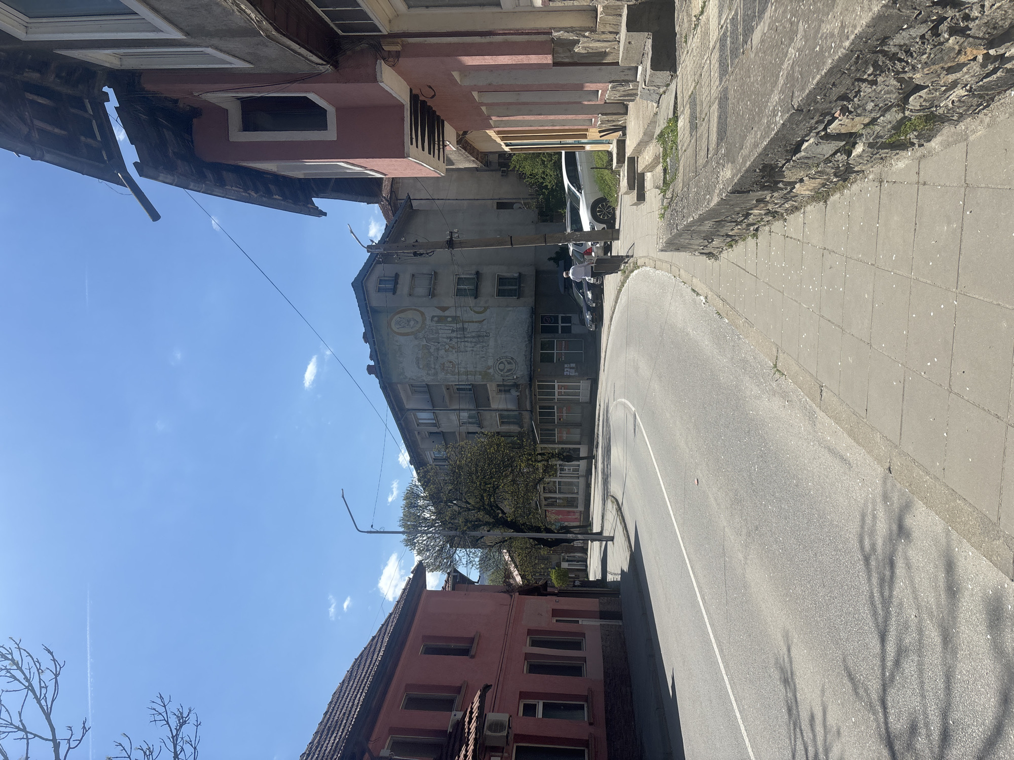 The image captures a street in Troyan, Bulgaria, featuring an asphalt road with a painted white line curving between buildings and a raised sidewalk. Buildings of varied architecture line the street. A faded mural adorns one of the buildings. Two individuals appear to be standing next to parked cars. A power pole is visible along the street. Tree shadows cast on the street are apparent. The sky is blue with some clouds.