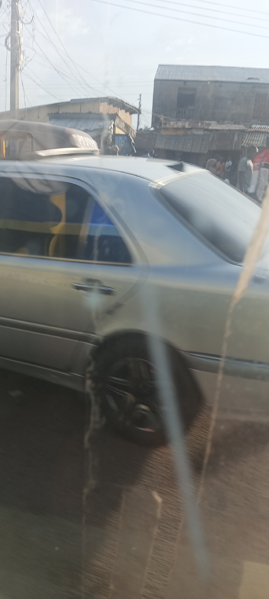 A slightly blurred, eye-level shot captures a silver or light-colored sedan car in motion on what appears to be a street in Gura Top, Nigeria, during the daytime. The car, possibly a Mercedes-Benz C-Class (W202 or W203 generation), is seen from its rear-left side, showcasing its body lines, tinted rear windows, and aftermarket black multi-spoke wheels.
The image seems to be taken from inside another vehicle or through a window, indicated by foreground blur, reflections, and vertical smudges. In the background, typical urban or semi-urban architecture is visible, including buildings with corrugated metal roofs and numerous overhead power lines strung between poles. Several people are discernible in the background, though they are heavily blurred, suggesting a bustling environment or street activity.
The sky is bright and clear, indicating sunny weather. On the roof of a vehicle (partially obscured) in the immediate foreground, faint white lettering on a darker surface can be seen, which appears to read "PT S09" or "PT 509".