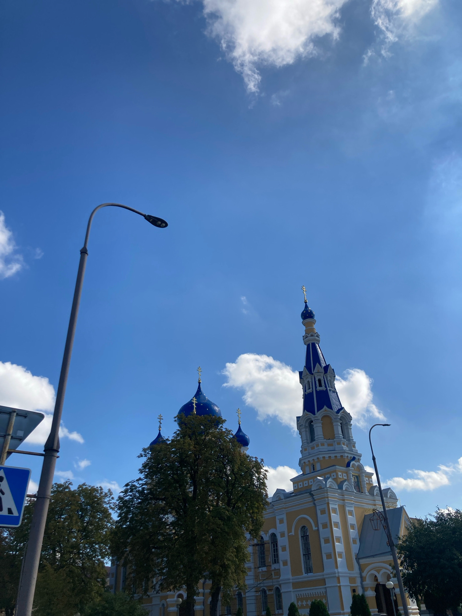 A bright, clear day in Brest, Belarus, is captured with a low-angle shot featuring a street light in the foreground on the left, and a yellow church with blue domes and spires on the right. The church's intricate architecture is visible, with multiple windows and decorative elements. Trees with green foliage are situated in front of the church, partially obscuring its lower section. A pedestrian crossing sign is mounted on a pole to the left of the street light. The sky is a vibrant blue with scattered white clouds, and sunlight casts shadows, indicating the time is likely mid-day. The overall scene depicts a serene urban landscape.