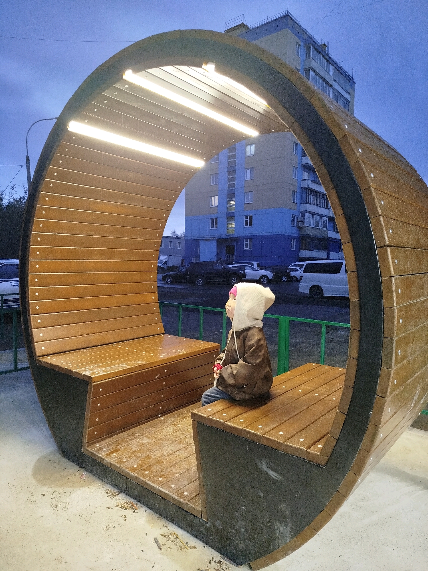 A young child wearing a brown jacket and a white hood sits on a wooden bench inside a large, curved wooden structure with built-in lighting. The structure appears to be a modern seating area or shelter. The background shows a multi-story building with blue and beige facades, and several cars parked on the street. The scene is outdoors, likely in a residential area or public space, and the lighting suggests it is either dusk or a cloudy day. The ground is paved, and there is a green metal fence in the foreground.