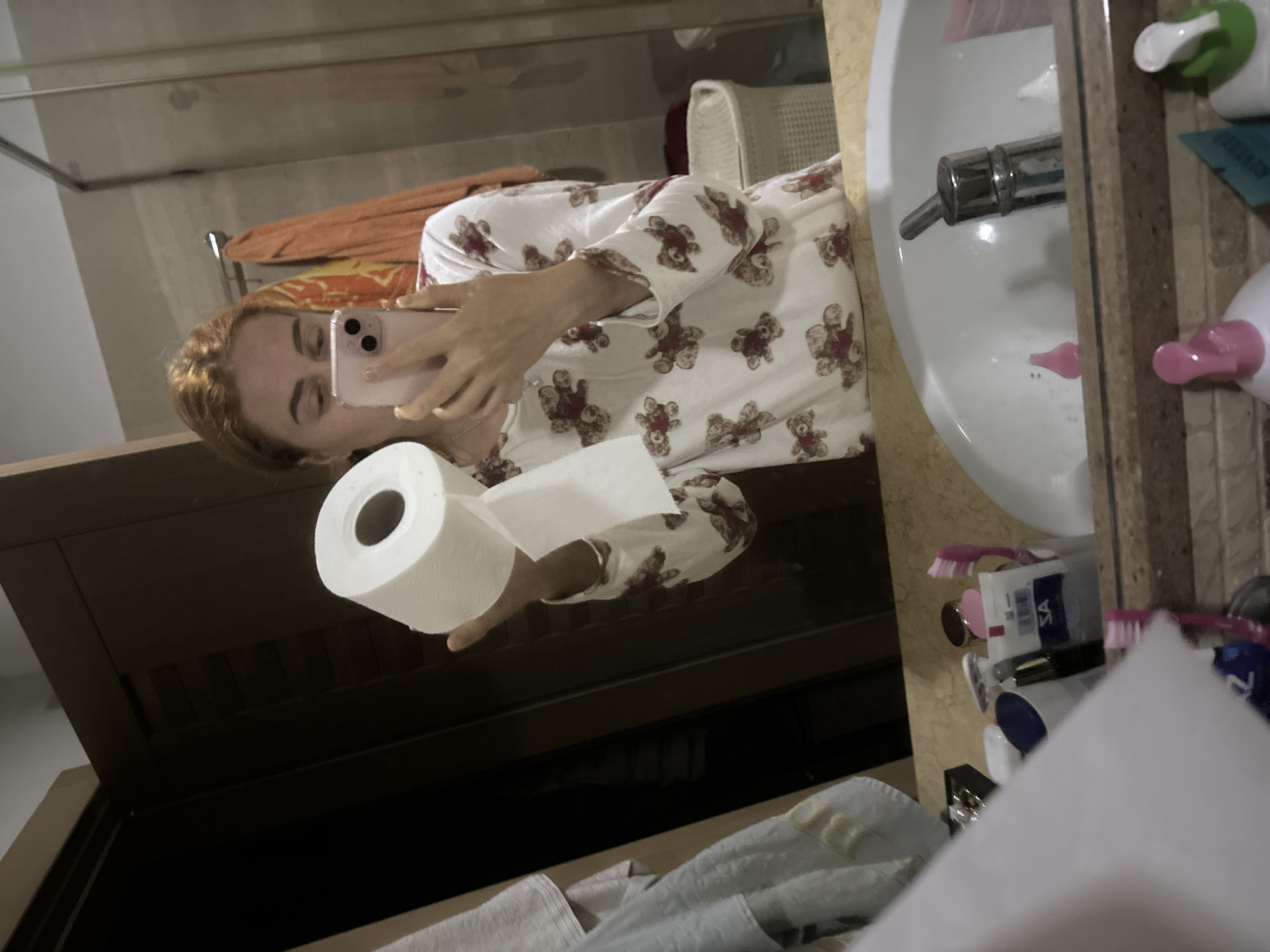 The image depicts a woman in a bathroom, taking a selfie in a mirror. She is wearing a white pajama top with a teddy bear print and holding a roll of toilet paper in her left hand. The bathroom features a sink with a chrome faucet, various toiletries, and a cabinet. There is an orange towel hanging on a rack in the background. Although the image lacks explicit location details, it is potentially set in Budva, Montenegro as requested, but the image itself provides no data to confirm.