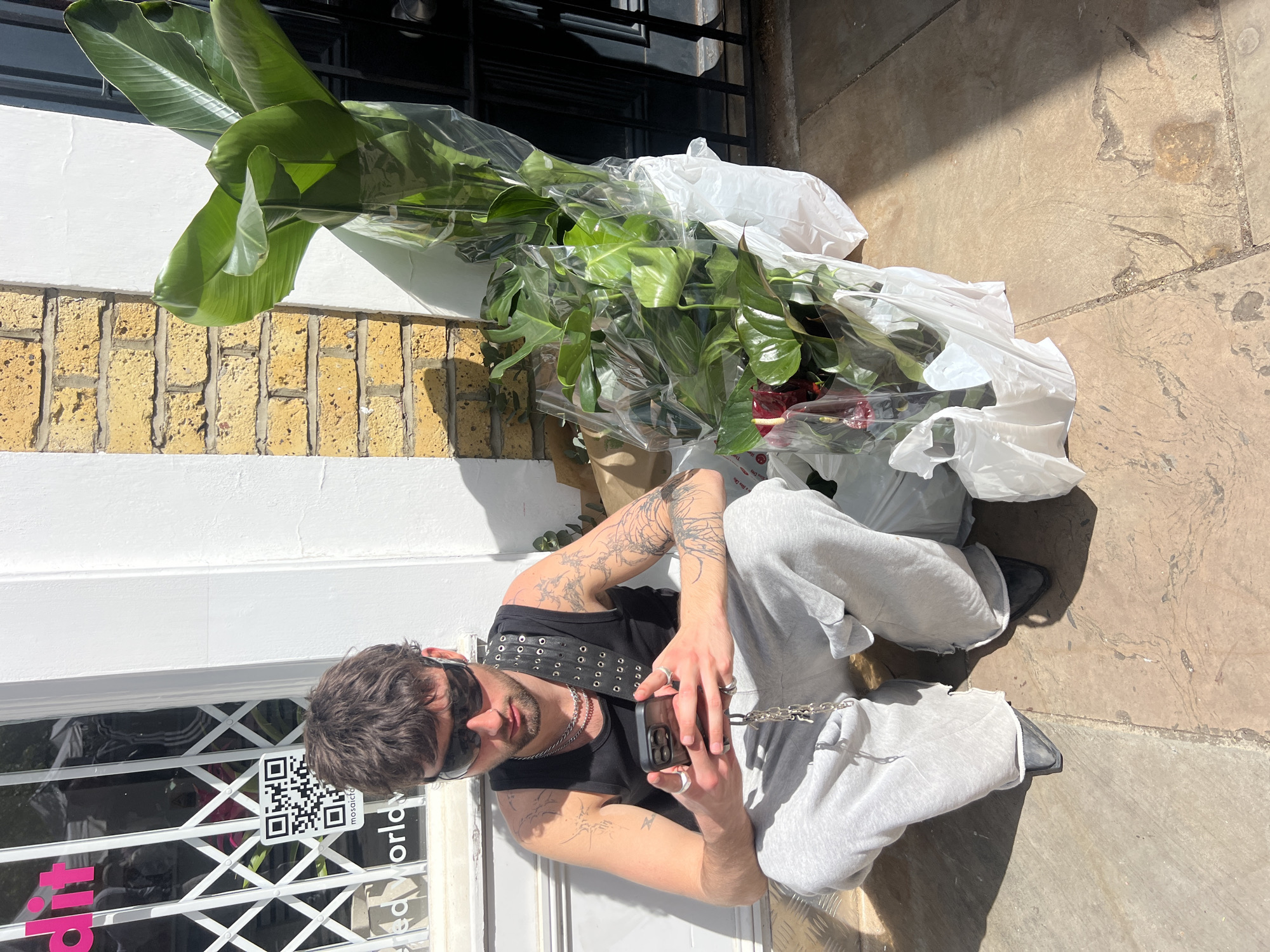 A man wearing sunglasses, a black tank top, grey sweatpants, and black shoes is sitting on a white doorstep next to a closed shop with metal bars and a pink sign on the window. A QR code sticker is visible on the window. The man is holding a smartphone and appears to be taking a photo. Next to him, leaning against a brick wall, are several large, wrapped plants. The ground is paved stone, and the location appears to be in London, United Kingdom.