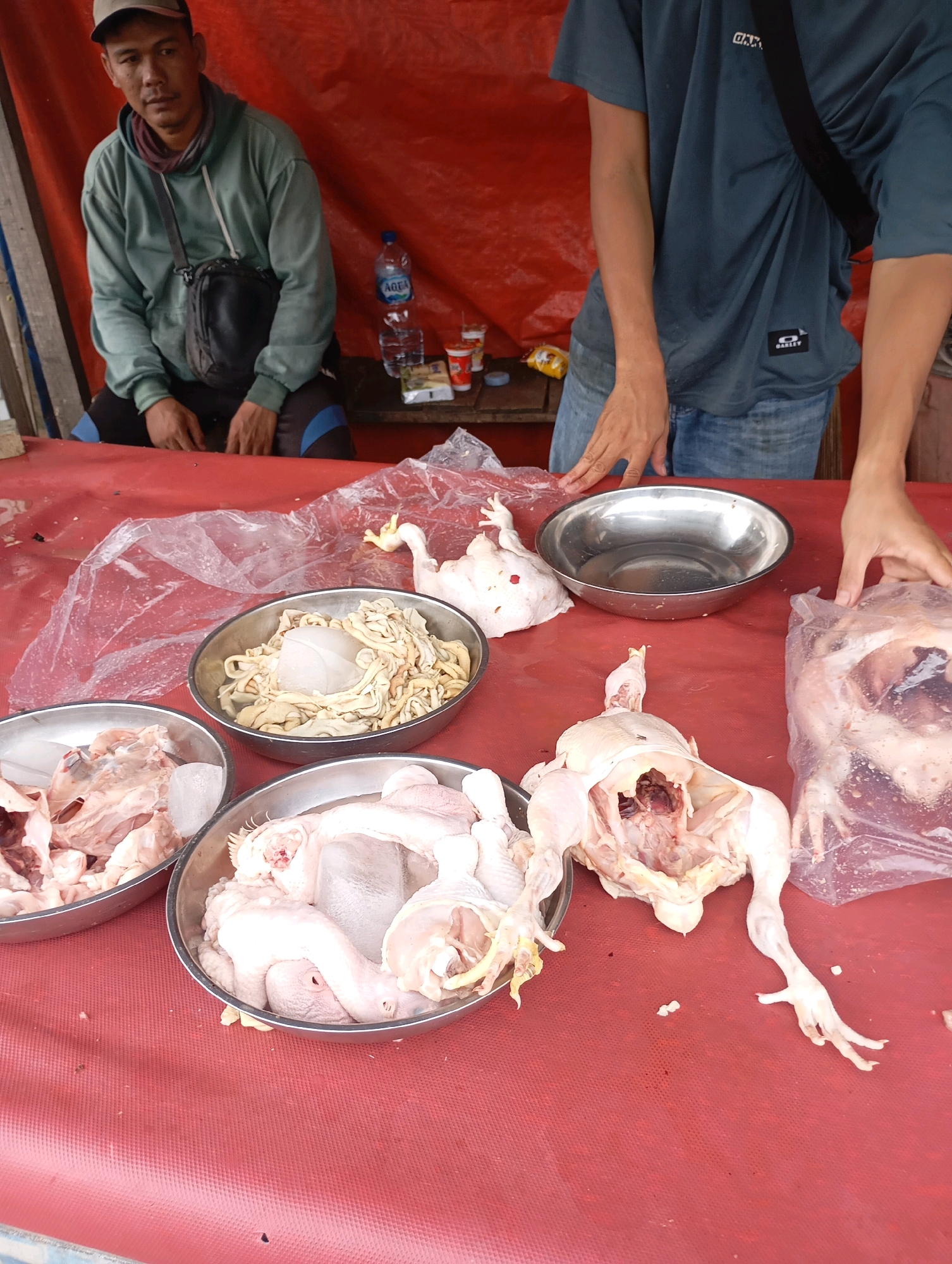 The image depicts a market stall in Babelan, Indonesia, where a vendor is selling raw chicken. A man wearing a green hoodie and a cap is seated behind a red table, and another person, partially visible, stands to the right. Several bowls of raw chicken parts, including whole chickens and intestines, are arranged on the table. One chicken is laid out on the table, gutted and prepared for sale. Another whole chicken is bagged and ready for purchase. The stall is set up with a red tarpaulin as a backdrop. The overall scene suggests a typical day at a local Indonesian market.