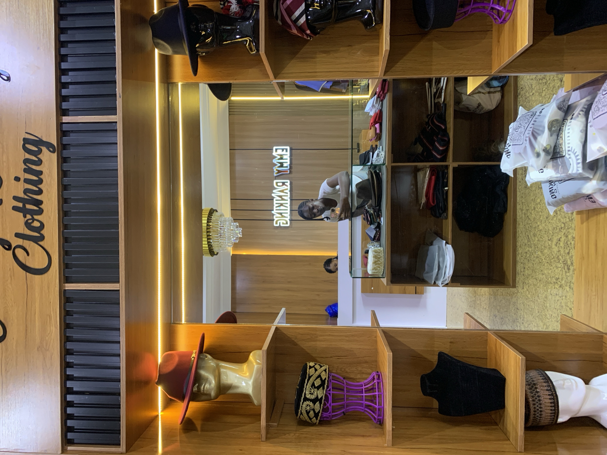 This is a photograph of the interior of a retail clothing store, likely in Gura Top, Nigeria, as indicated by the signage. The store features wooden shelving units displaying various apparel and accessories, primarily hats. On the left side, a mannequin head wears a red fedora. Below that, a purple stand displays a traditional cap. Further down, another mannequin head, this one white, wears a patterned cap. The right side of the store also has shelving with more hats on mannequin heads, as well as folded garments. In the background, a mirrored wall reflects the store's interior, showing a woman standing behind a counter, seemingly interacting with a mobile phone. A neon sign on the mirrored wall reads "EMMY RANKING" in reverse. The lighting in the store appears to be a combination of overhead and strip lighting, creating a warm ambiance. The floor is tiled in a yellowish, textured pattern. The overall impression is that of a well-stocked boutique specializing in men's headwear and clothing.