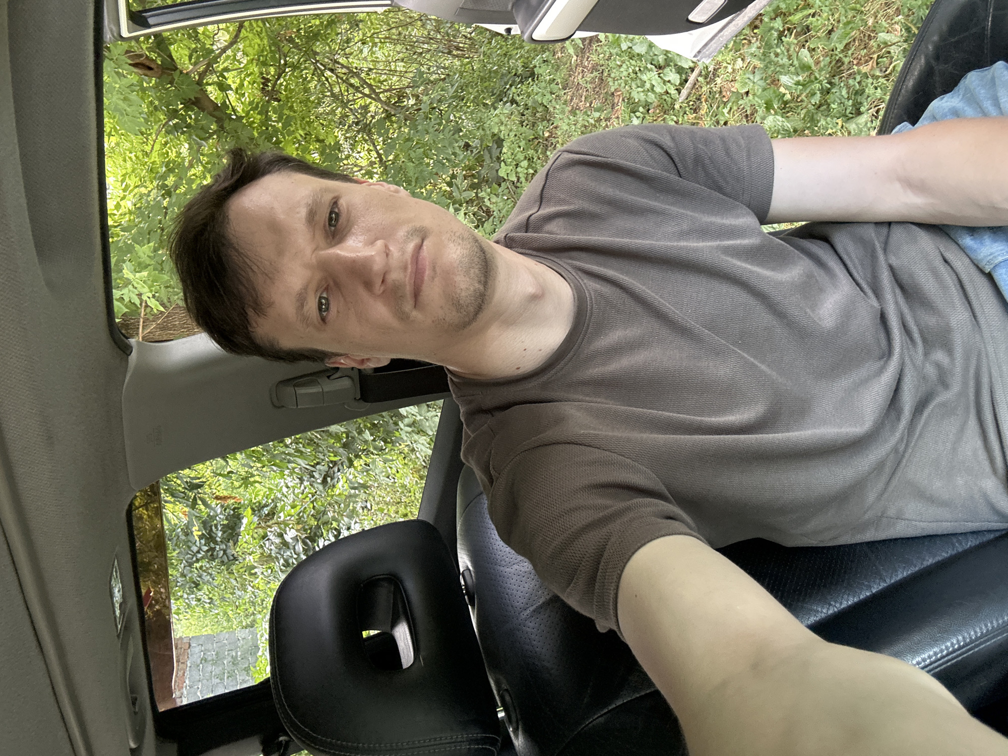 In the city of Makiv, Ukraine, a man with short brown hair and a neutral expression sits in the driver's seat of a car. He is wearing a brown, short-sleeved t-shirt and blue jeans. The car's interior is visible, including a black leather headrest and seat. Through the open car door and windows, lush green foliage and trees are visible, suggesting an outdoor, possibly rural or suburban setting. The lighting indicates it is daytime.