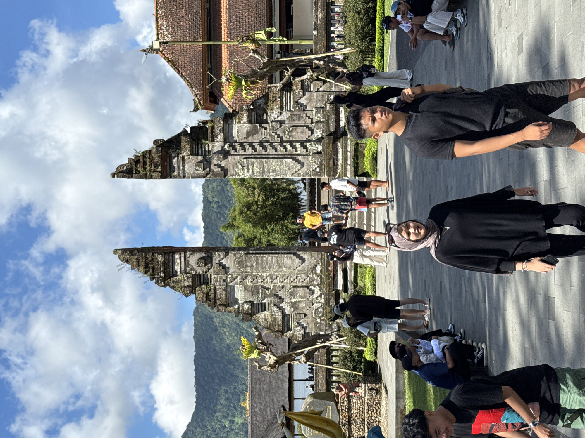 The image shows the Handara Gate in Baturiti, Indonesia. The gate is a stone structure, with mountains visible in the background. There are people on the paving stone area surrounding the gate.