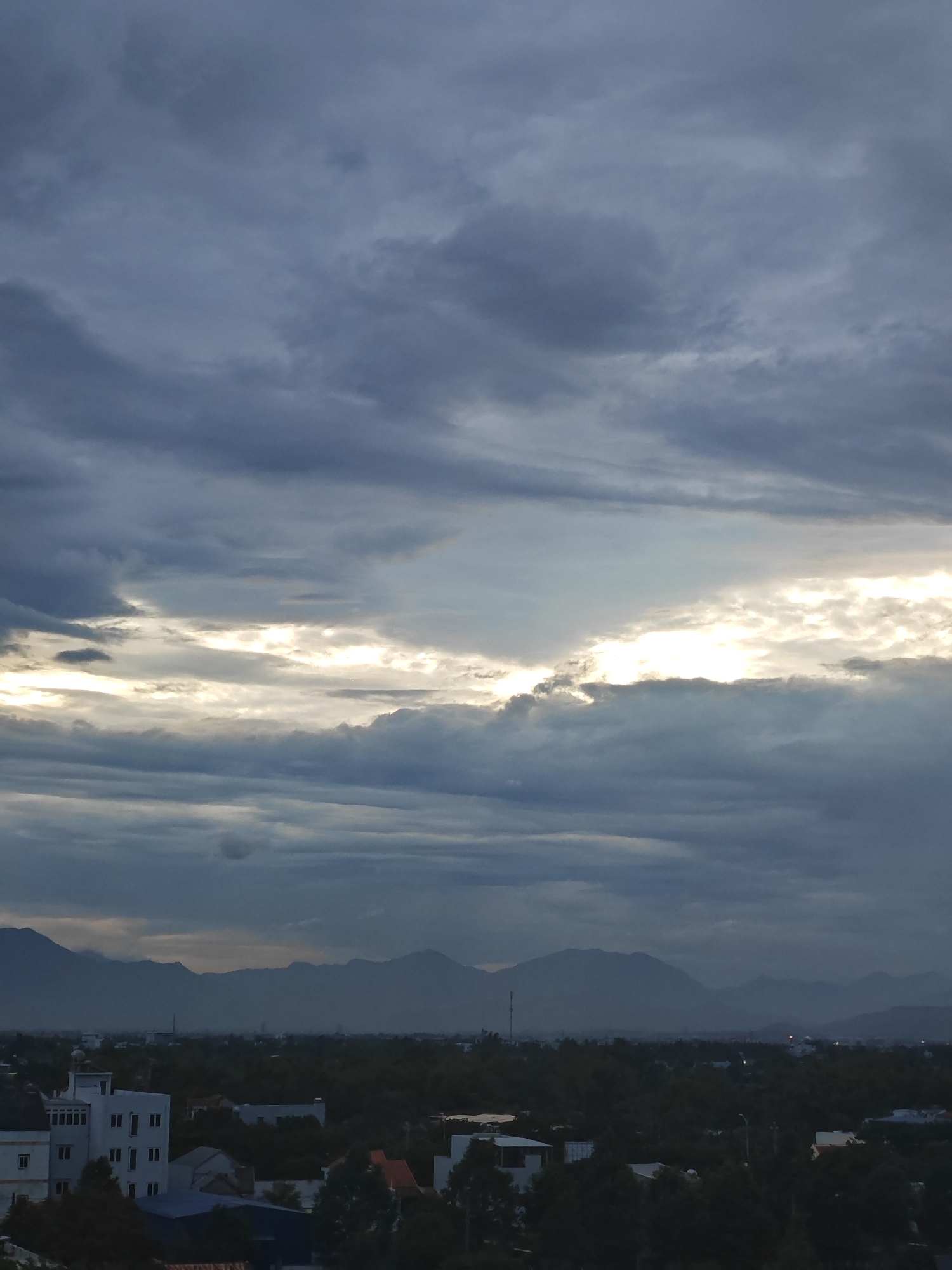 This is a vertical, eye-level, outdoor, daytime, full shot of a cloudy sky over a city and mountains. The sky is filled with heavy, dark gray clouds, but there is a break in the clouds where bright, yellowish-white light is visible. The lower half of the image shows a city skyline with trees and buildings, and a mountain range in the distance. The city appears to be located in Da Nang, Vietnam, based on the characteristic mountainous backdrop and the architecture of the buildings. The overall atmosphere is one of a dramatic and overcast sky, likely before or after a storm. There are no people or animals visible in the image.