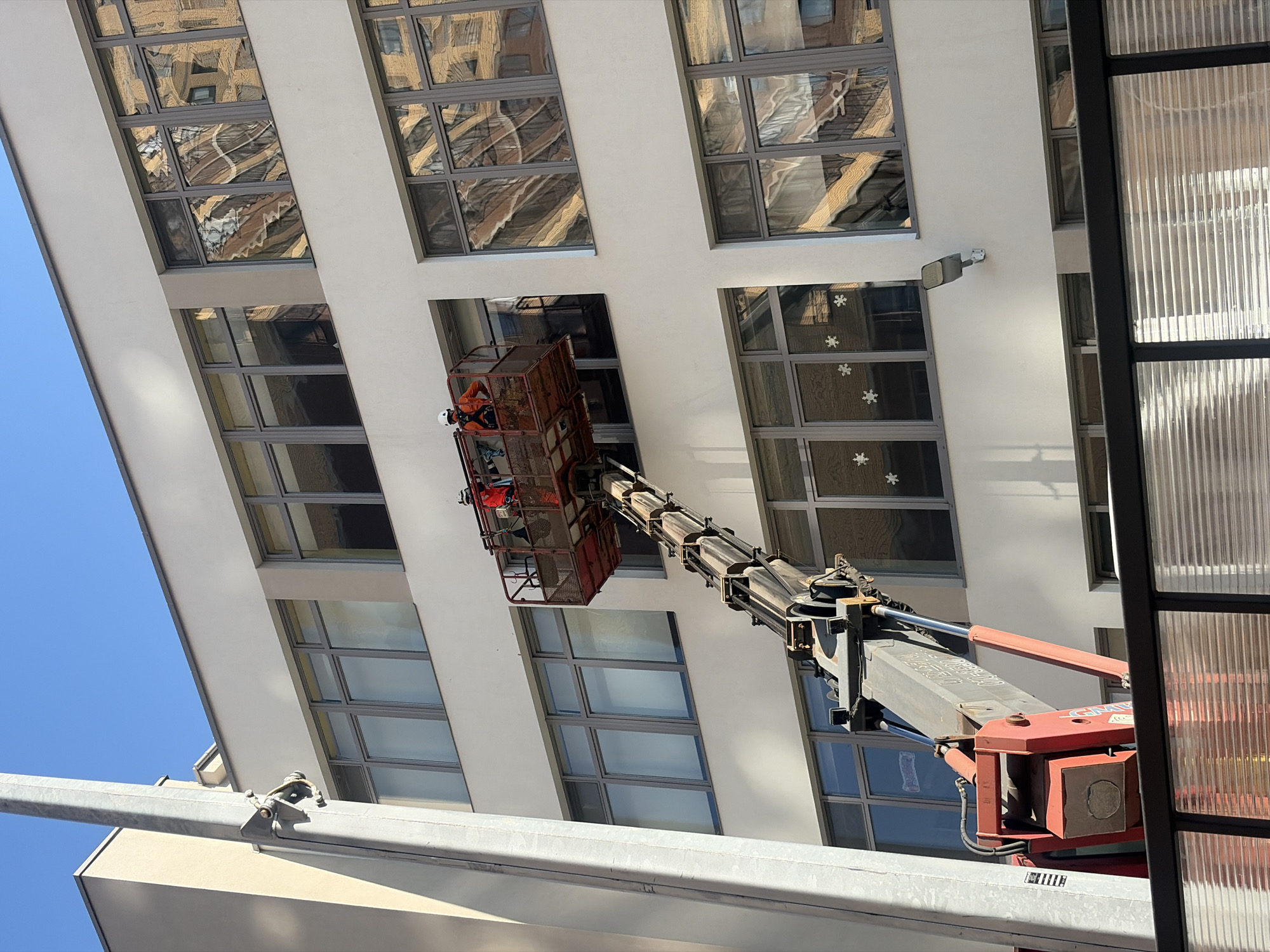 A low-angle, medium shot shows a cherry picker extending towards a building with many windows in Saint Petersburg, Russia. The sky is clear and blue. Two workers wearing orange safety vests and white helmets are in the cherry picker's basket. The building has a white facade with gray-framed windows. Some windows have snowflake stickers. The cherry picker has a red basket and gray extendable arm. The windows reflect images of other buildings. The cherry picker is partially obstructed by a frame-like object with a translucent, ribbed panel.