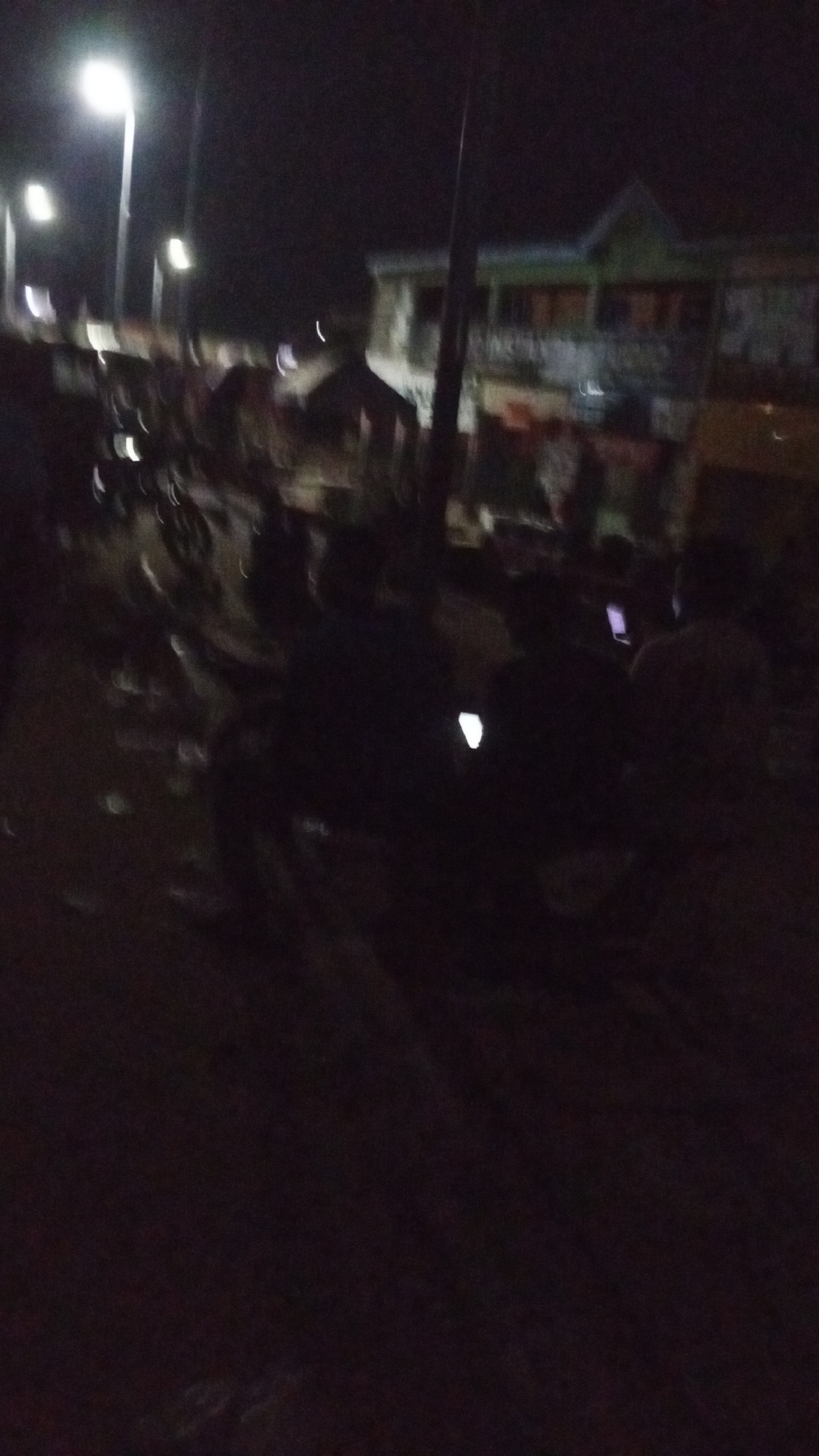 The image captures a dark, blurry night scene in what appears to be a street or public gathering area in Lokoja, Nigeria. The dominant features are several bright, out-of-focus streetlights illuminating the upper left and middle-left portions of the frame, casting a soft glow.
In the foreground and midground, numerous people are visible, mostly as dark silhouettes against the ambient light. They appear to be gathered, possibly sitting or standing. A notable detail is the presence of several glowing rectangles, clearly identifiable as phone screens, held by individuals in the midground, suggesting active engagement with mobile devices. The low light and motion blur make it difficult to discern facial features or specific details of clothing.
In the background, a multi-story building with a distinctive green roof and lighter-colored walls is visible, though also blurry. The architectural style seems typical of residential or commercial structures in an urban or semi-urban setting. What might be utility poles or other vertical structures also extend upwards into the dark sky. Suggestions of other vehicles or objects are present in the blurred foreground, but are indistinct. The overall impression is of a lively but dimly lit nighttime street scene where people are congregated, possibly socializing or waiting. No visible text can be deciphered.