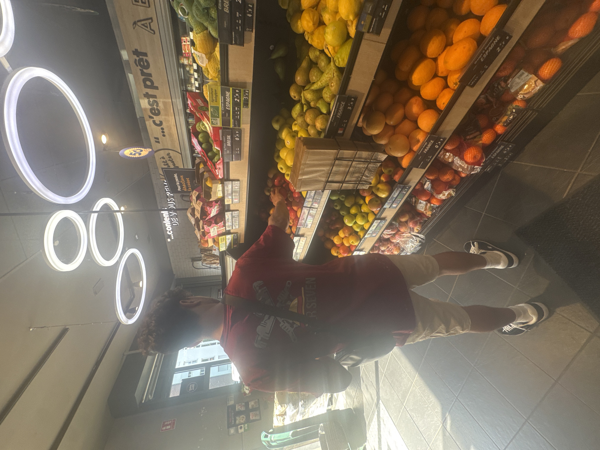 Here's a detailed description of the image:
**Scene:** The image captures a scene inside a supermarket, presumably in Marcq-en-Barœul, France, based on OCR data. The location appears to be the produce section.
**People:** A young man is standing in front of a display of fruits. He is wearing a red t-shirt with a design on the front, beige shorts, white socks, and black and white sneakers. He has dark curly hair. He is reaching towards the fruit display.
**Objects:**
* Displays of fruits (possibly oranges, lemons, limes, tangerines, and tomatoes) with price tags and labels indicating the origin of the produce (France, Spain, and Brazil).
* Supermarket shelving.
* Fluorescent ring lighting on the ceiling.
* A metal shopping cart in the lower part of the picture.
* Price tags and labels in French.
**Actions & Interactions:** The man appears to be selecting fruits from the display, possibly choosing which ones to purchase.
**Environment:** The store seems well-lit with tiled floors. There is a window in the background that indicates a building or street outside. The aisle is relatively clean.