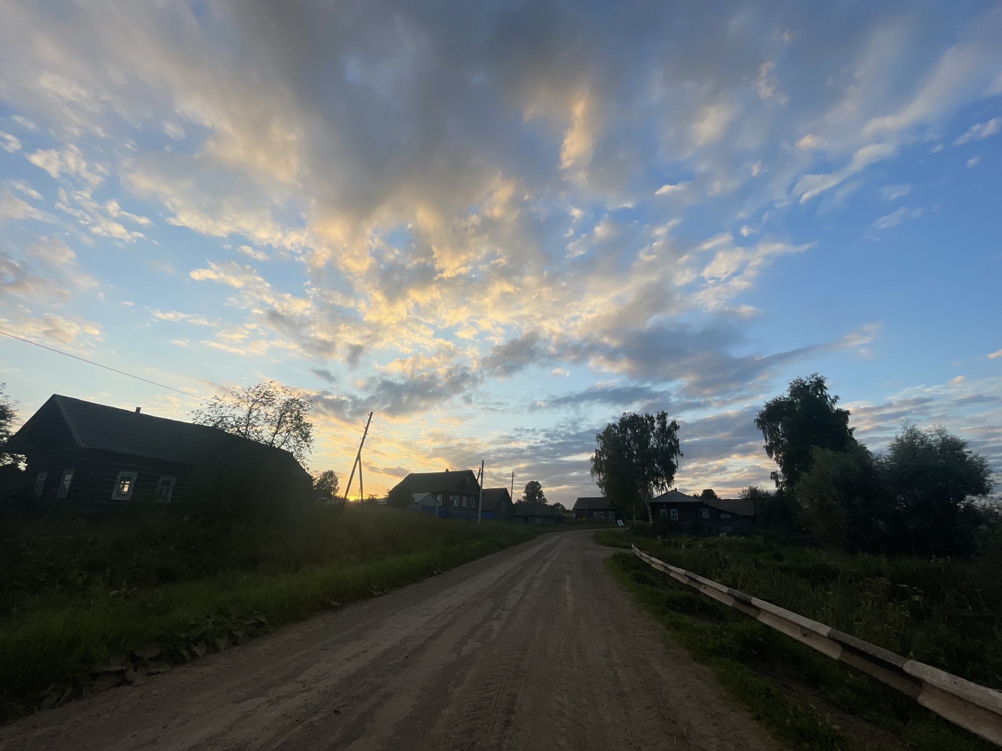 The image depicts a rural village scene in Panteleevka, Russia, at dusk. A dirt road curves into the distance, lined with wooden houses and lush green vegetation. The sky above is a canvas of blue, adorned with scattered clouds painted in hues of orange and yellow by the setting sun. A metal guardrail runs along the right side of the road, indicating its presence within a populated area. The overall atmosphere is serene and tranquil, characteristic of a peaceful countryside setting.