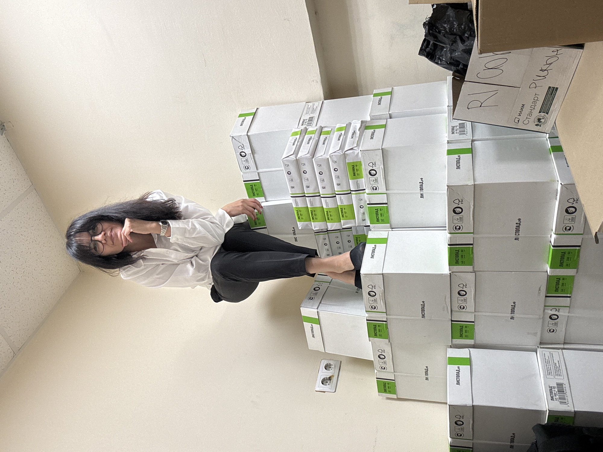 A medium shot shows a woman sitting on a stack of boxes in an indoor environment, possibly a storage room or an office space, in Unknown, Unknown. She is dressed in black trousers and a white button-down shirt, and she is wearing glasses. She leans on her hand with a contemplative expression. The boxes stacked next to her are white with green accents and appear to contain paper. The brand name "ЛИСТОПАД РФ" is printed on the boxes, along with other symbols and text. There is a power outlet near the base of the boxes. Other boxes are also visible in the upper right of the shot. The walls of the room are light beige.