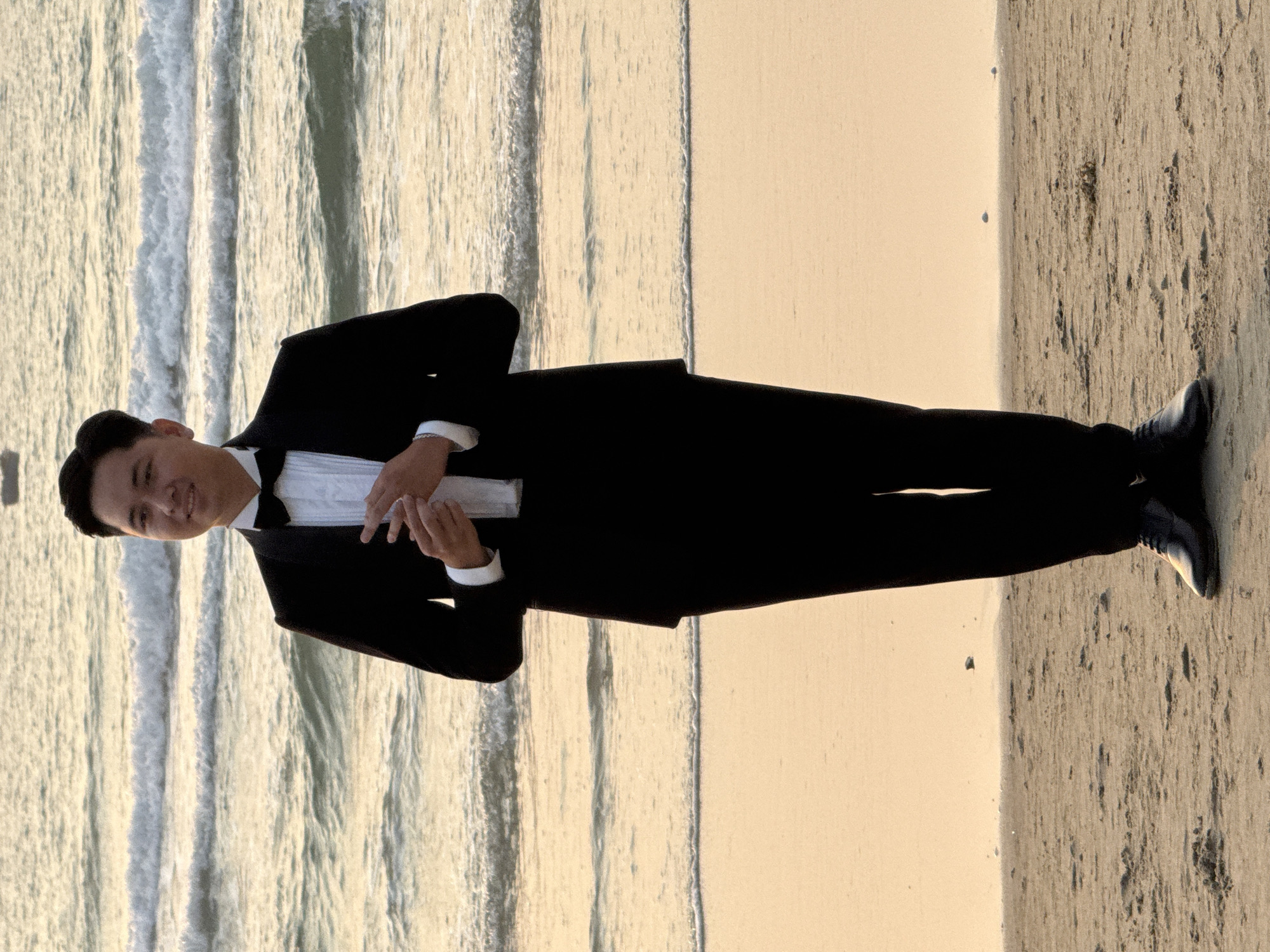 A man wearing a black tuxedo stands on a beach in Đà Nẵng, Vietnam. The man is positioned at an angle relative to the beach and the ocean, which is visible in the background. The man looks to be facing the viewer. The sand is beige and textured. The ocean is turbulent and contains waves.