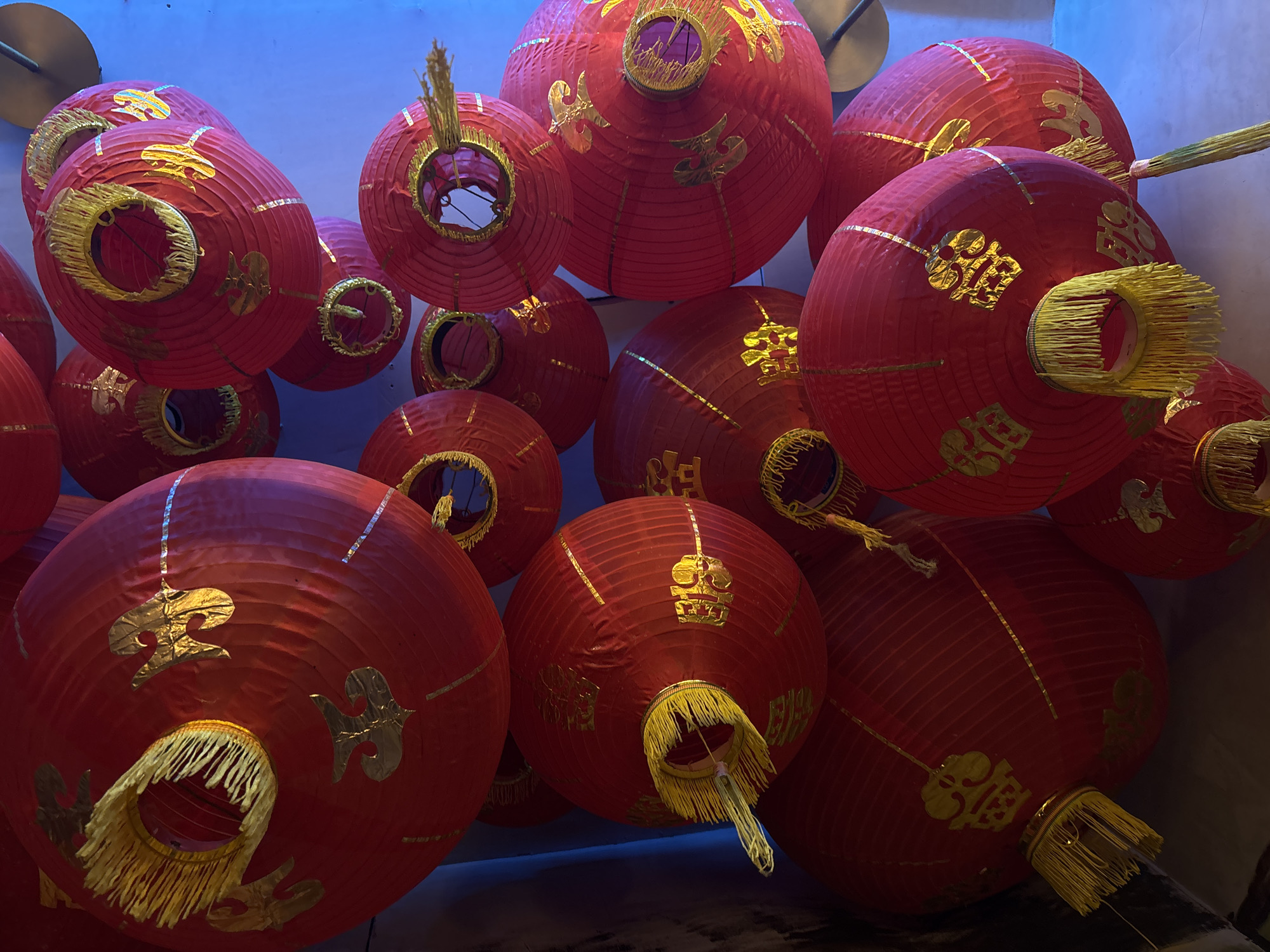 The image depicts a cluster of red Chinese lanterns, likely silk or paper, hanging indoors. The lanterns are adorned with gold-colored embellishments. The lanterns vary in size, and the arrangement creates a dense, overlapping composition. The lighting in the scene is somewhat dim, casting shadows and creating a slightly dramatic effect. The location is inferred to be in North Kuta, Indonesia.