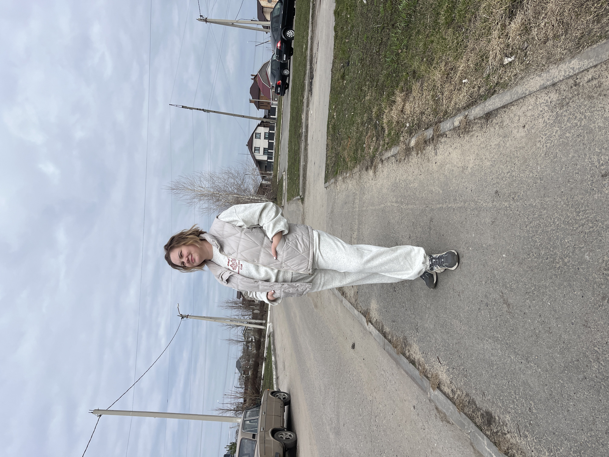A young woman stands on a sidewalk in Usman, Russia, based on the cars and buildings. She is wearing a light-colored sweat suit and vest with dark athletic shoes. She has her hand in her vest pocket. The sidewalk is adjacent to a street. Several houses and cars can be seen in the background. Utility poles and wires are overhead. The sky is overcast.