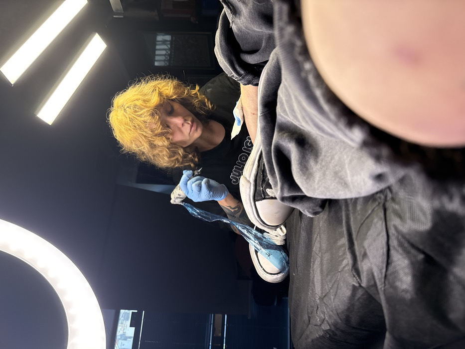A person with curly blonde hair is tattooing a client. The tattoo artist wears a black shirt and blue gloves. The client, positioned overhead, wears a gray hoodie and black and white sneakers. A tattoo machine wrapped in blue plastic is in use. Overhead lights and a ring light illuminate the scene.