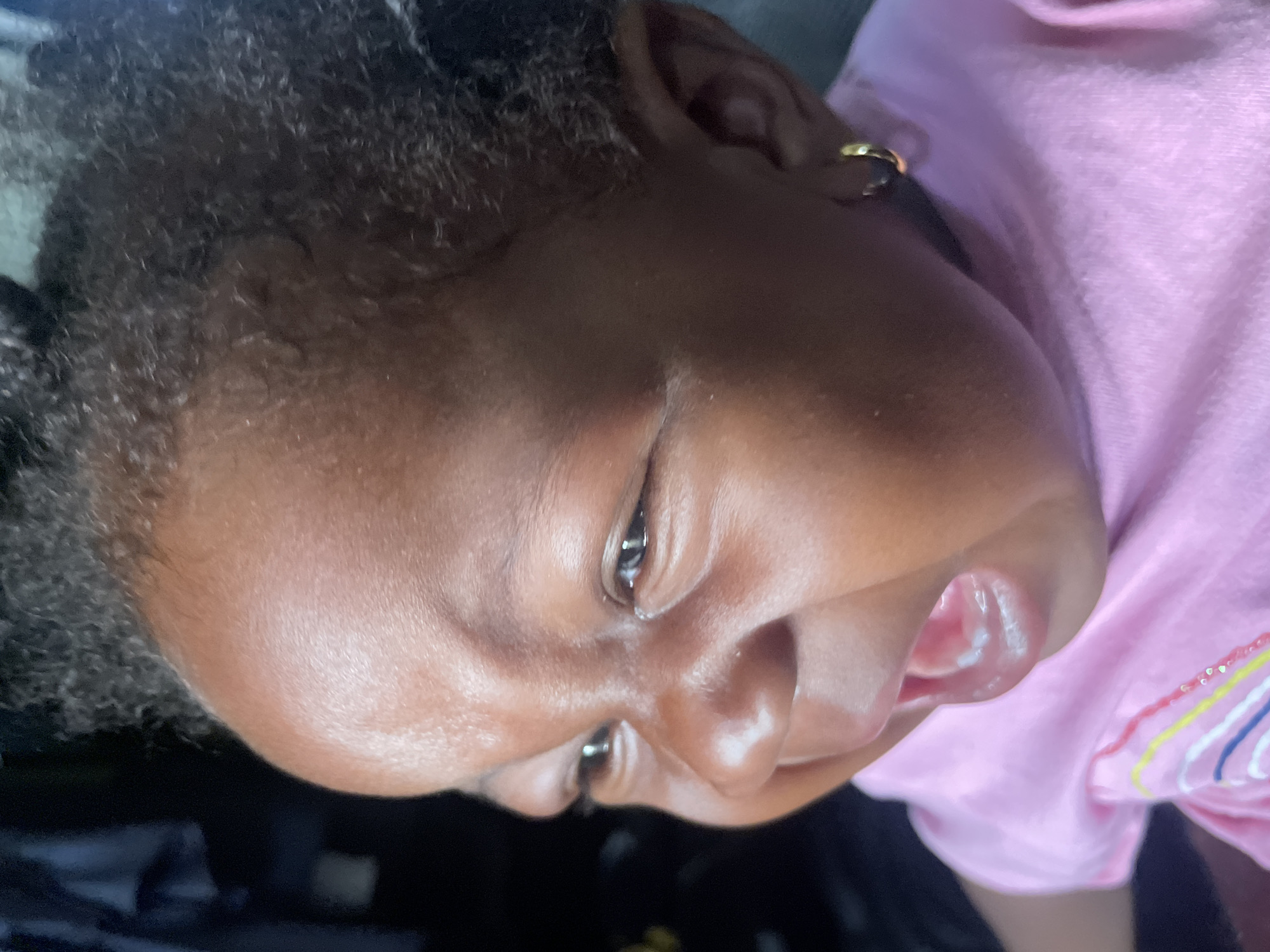A close-up, slightly out-of-focus image shows the face of a young Black child, likely a baby or toddler. The child has dark, curly hair and a gold earring in their left ear. They are wearing a pink shirt with a rainbow design. The child's mouth is slightly open. The location is not discernible from the image, and there is no indication it was taken in Lokoja, Nigeria.
