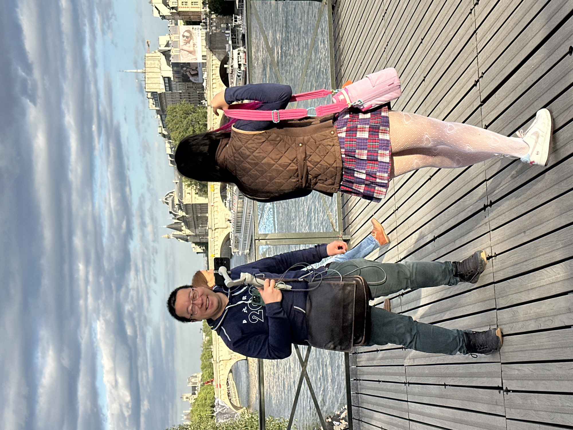 The image shows two people standing on a wooden bridge in Paris, France. One person is a man with glasses, wearing a navy blue hoodie, green pants, and black boots, carrying a black bag and what appears to be medical equipment. He is smiling at the camera. The other person is a woman with long black hair, wearing a brown quilted jacket, a plaid skirt, heart-patterned white tights, and white sneakers, and carrying a pink bag. The backdrop includes a view of the Seine River, Parisian buildings, and the sky with clouds. The composition is slightly tilted.