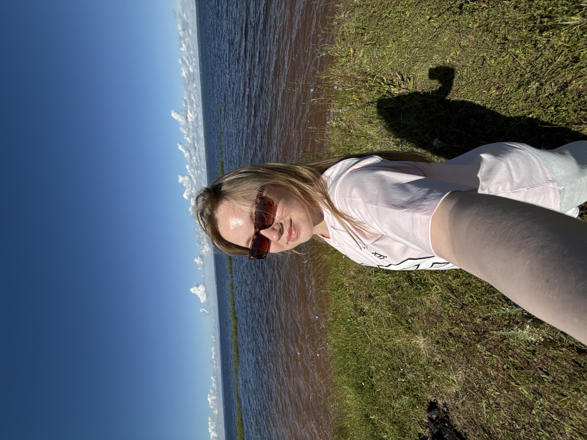 A young woman with blonde hair and sunglasses is taking a selfie on a grassy area near a large body of water. She is wearing a pink t-shirt and light blue pants. The sky is clear and blue with some scattered white clouds. The water is a reddish-brown color and appears to be a lake or river. The area is sunny and appears to be a natural landscape.