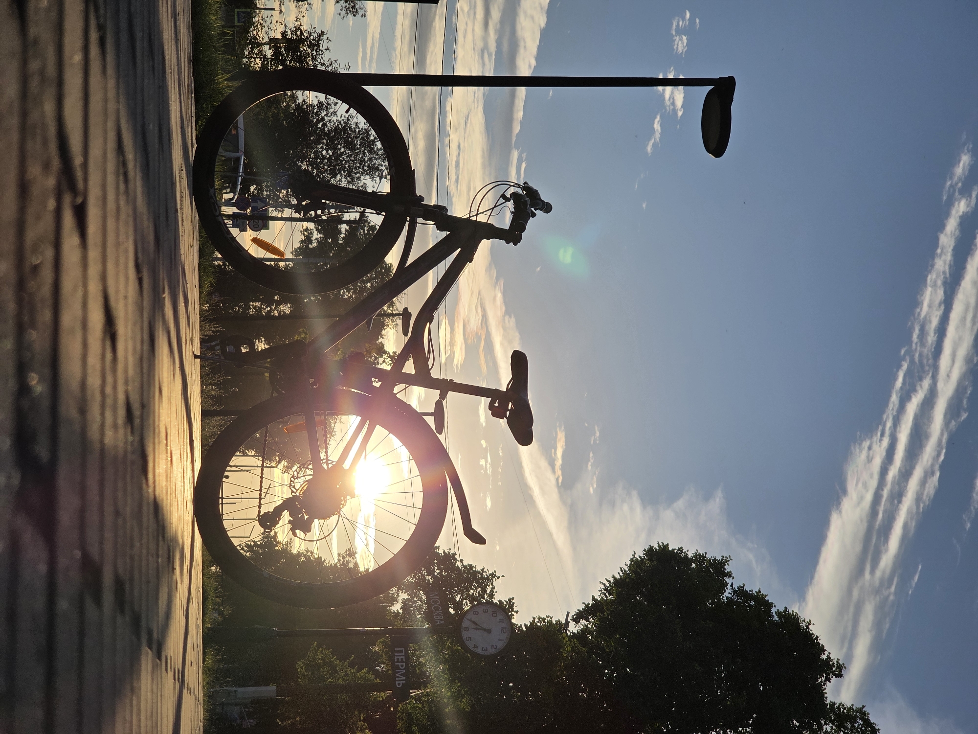 A bicycle is silhouetted against a sunset sky, its frame and wheels outlined by the golden light. The bicycle is parked on a brick pathway, with its handlebars facing towards the right side of the frame. A lamppost stands tall behind the bicycle, also silhouetted. The sky is a vibrant blue, streaked with wispy white clouds that catch the last rays of the setting sun. Trees, also silhouetted, form a dark band across the lower portion of the image, obscuring the ground behind the pathway.
In the foreground, the texture of the brick pathway is visible, showing individual bricks and the lines between them. The sun is low on the horizon, casting long shadows and creating lens flares that streak across the image. These flares give the scene a warm, ethereal quality.
A clock is visible attached to a pole, partially obscured by trees. The clock face shows the time to be approximately 7:00. Two signs are also visible, one with Cyrillic text that appears to say "ПЕРМЬ" (Perm), and another with text that appears to say "МОСКВА" (Moscow). The presence of these signs, combined with the context of Chaikovsky, Russia, suggests this is likely a public space within the city, possibly a park or a street.
The overall mood of the image is peaceful and serene, evoked by the beautiful sunset and the quiet stillness of the parked bicycle. The combination of natural elements like the sky and trees with man-made objects like the bicycle, lamppost, and clock creates a sense of urban nature. The time of day is clearly sunset, and the weather appears to be clear with some scattered clouds.