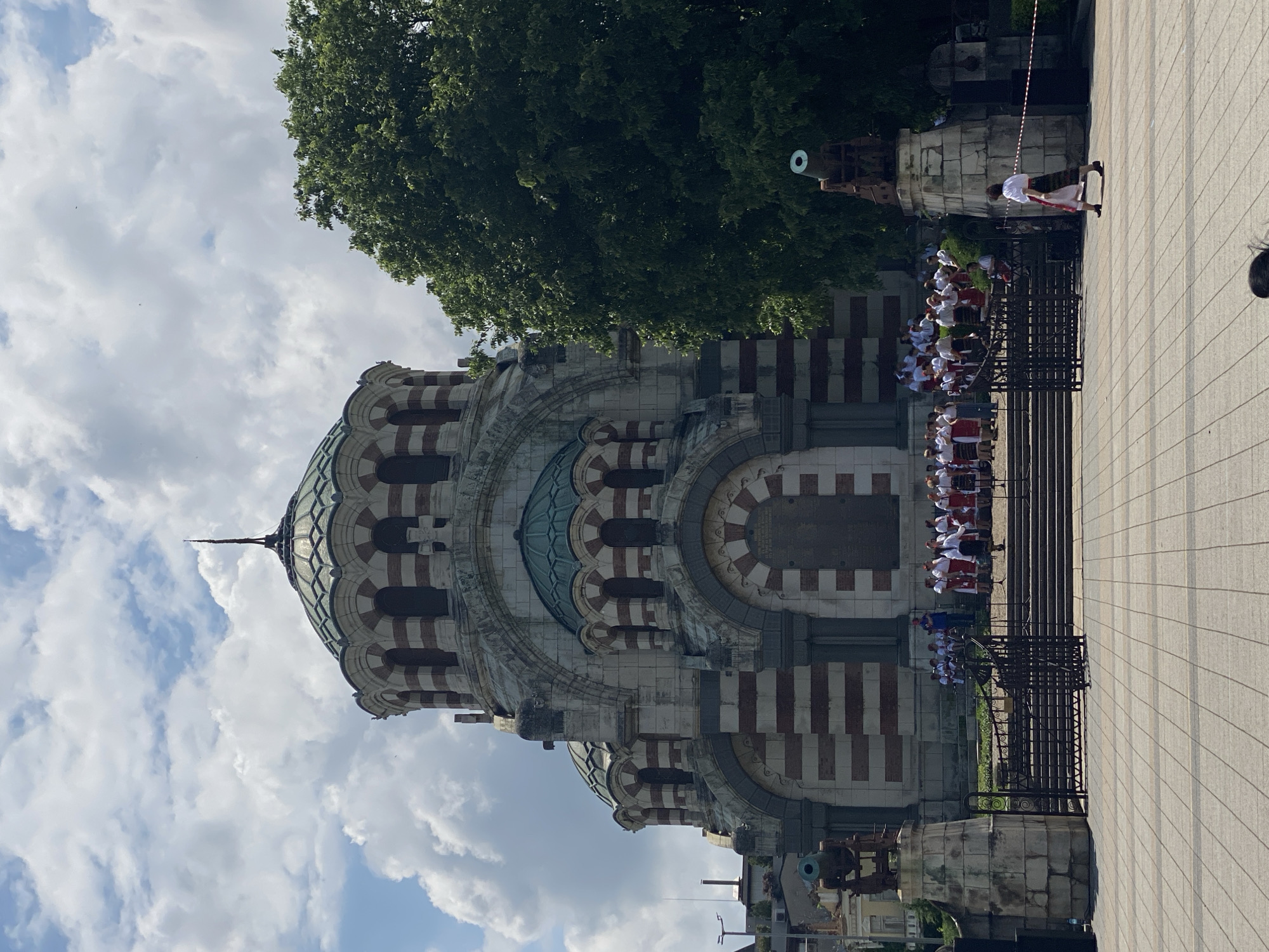 The image shows the exterior of the St. George the Conqueror Mausoleum Church in Pleven, Bulgaria. The church features a distinctive design with alternating white and red brick patterns on its dome and walls. A large tree is visible behind and to the side of the structure. In front of the church, a group of people dressed in what appear to be traditional Bulgarian costumes are lined up. A black wrought iron fence separates the group from a paved area. A cannon is visible to the side, positioned on a stone base. A woman in traditional Bulgarian dress is standing nearby. The sky is partly cloudy, suggesting daytime conditions.